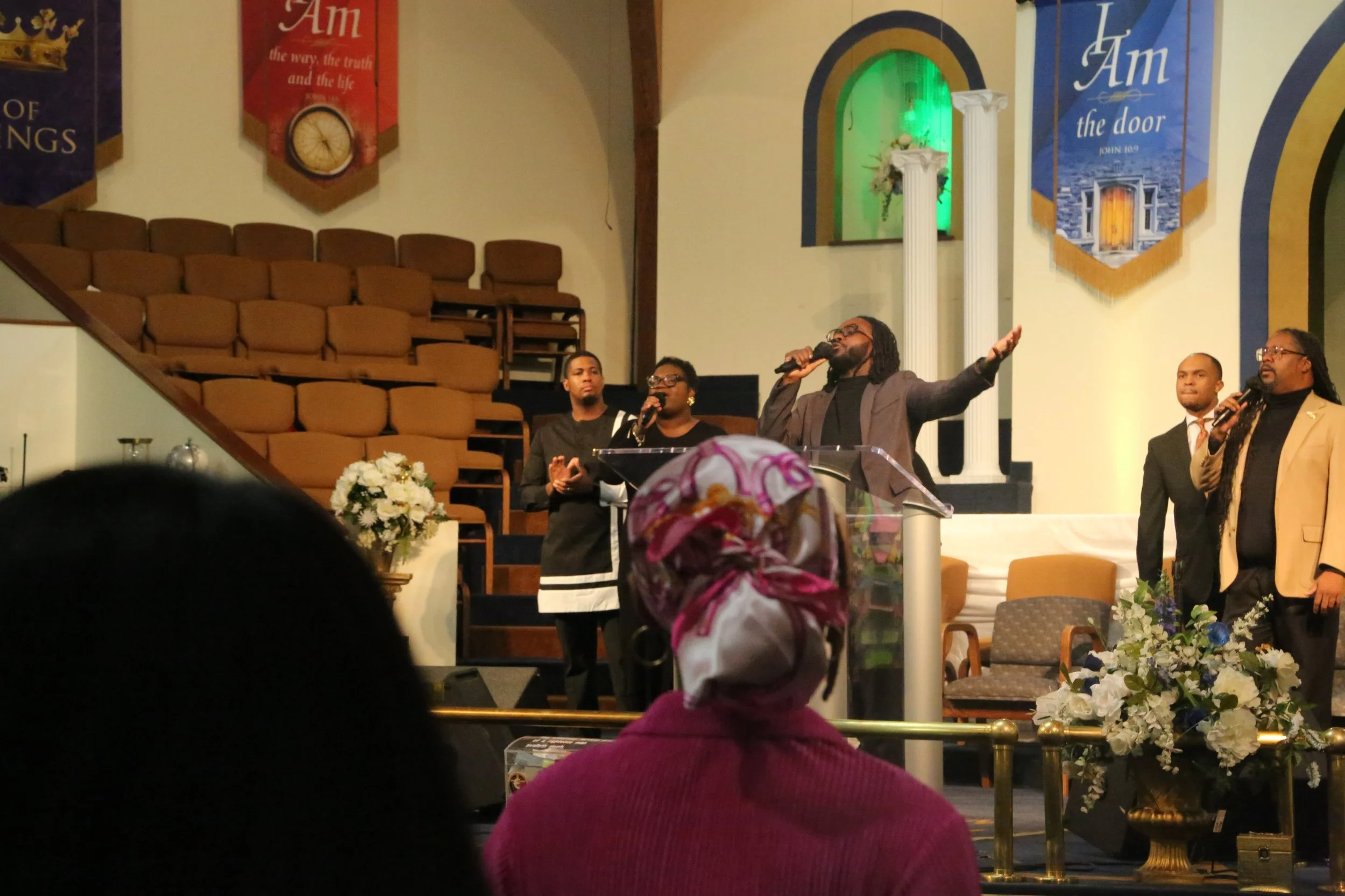 Church service with a preacher and choir singing on stage, congregation seated in the pews, floral arrangements, banners reading 'I Am' and 'I Am the door' from John 10:9 in the background.