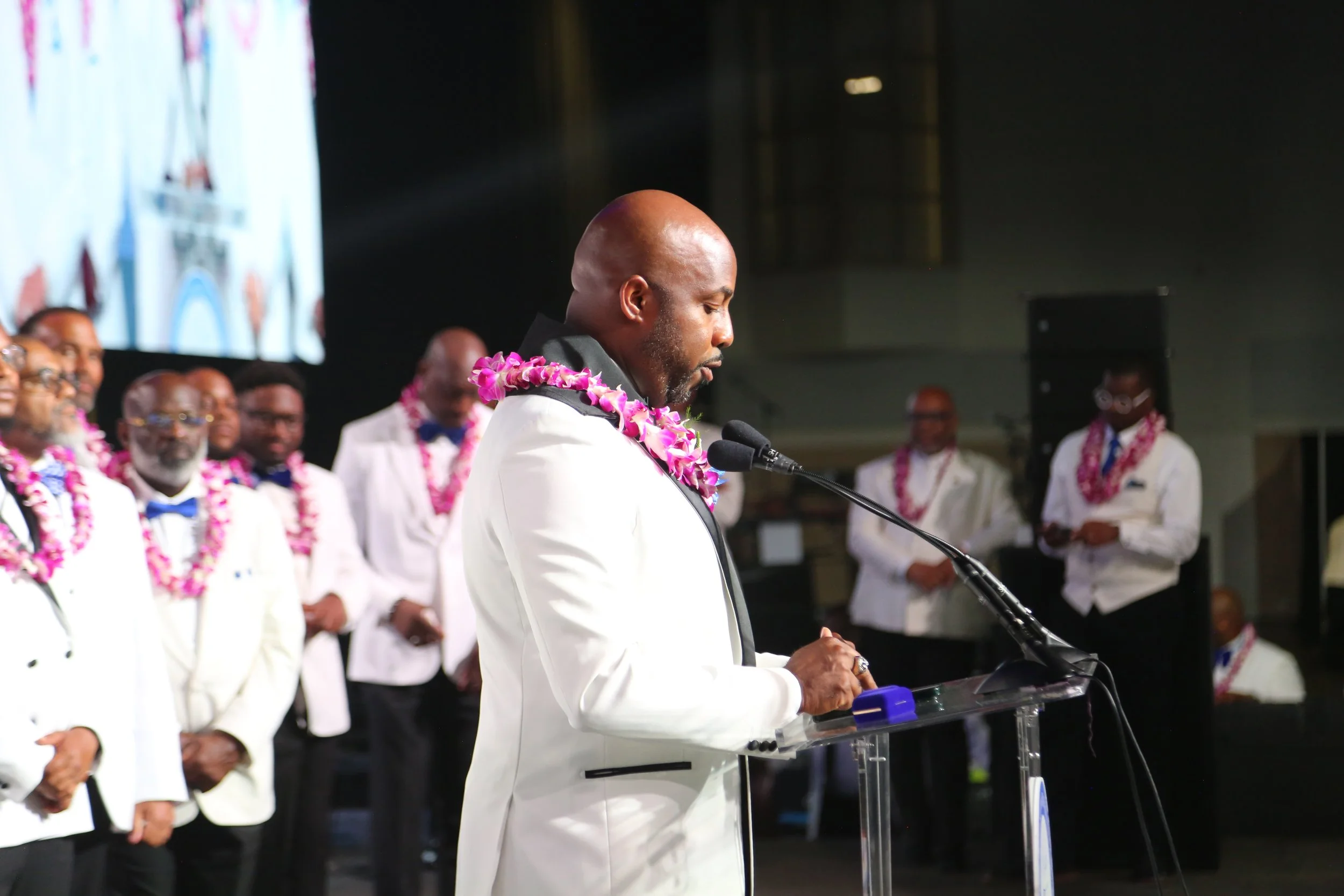 A man in a white suit with a purple lei stands at a podium, speaking into a microphone during a formal event. Several men in white suits with leis stand in the background.