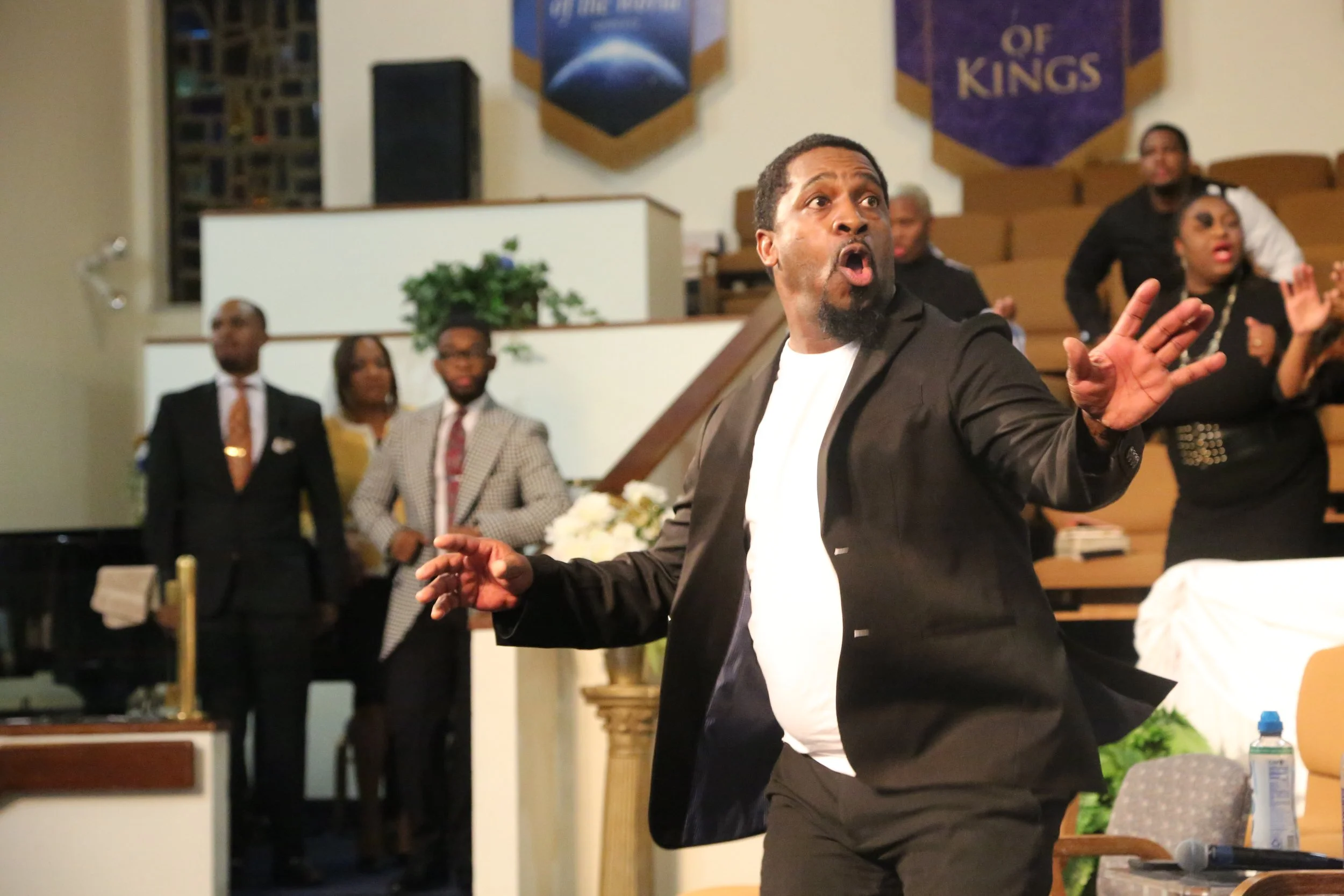 A man in a black suit speaking passionately in a church or religious setting, with several people standing and clapping in the background.