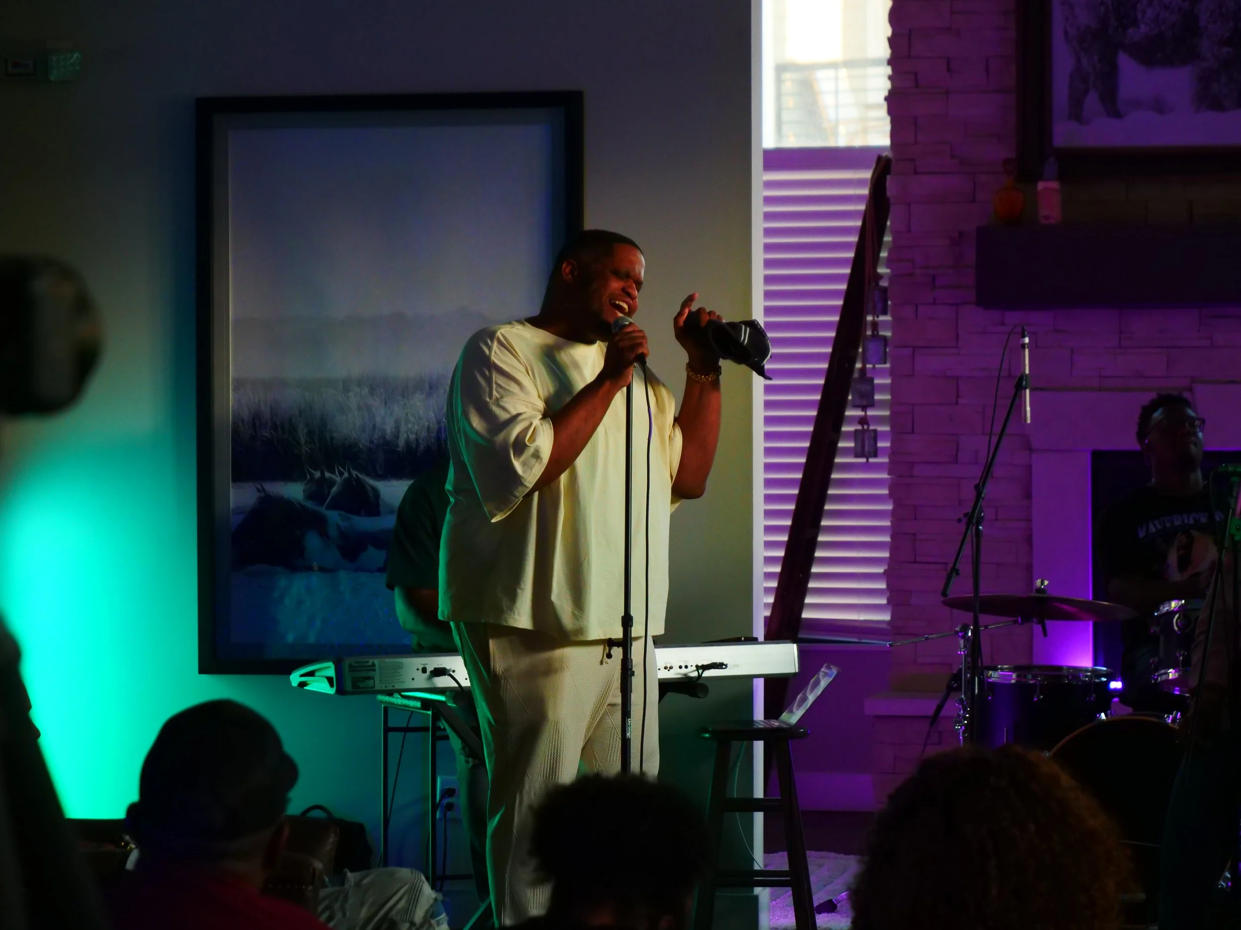 A man singing into a microphone on stage, holding a baseball cap, with a keyboard behind him and a drummer to the side in a dimly lit room.
