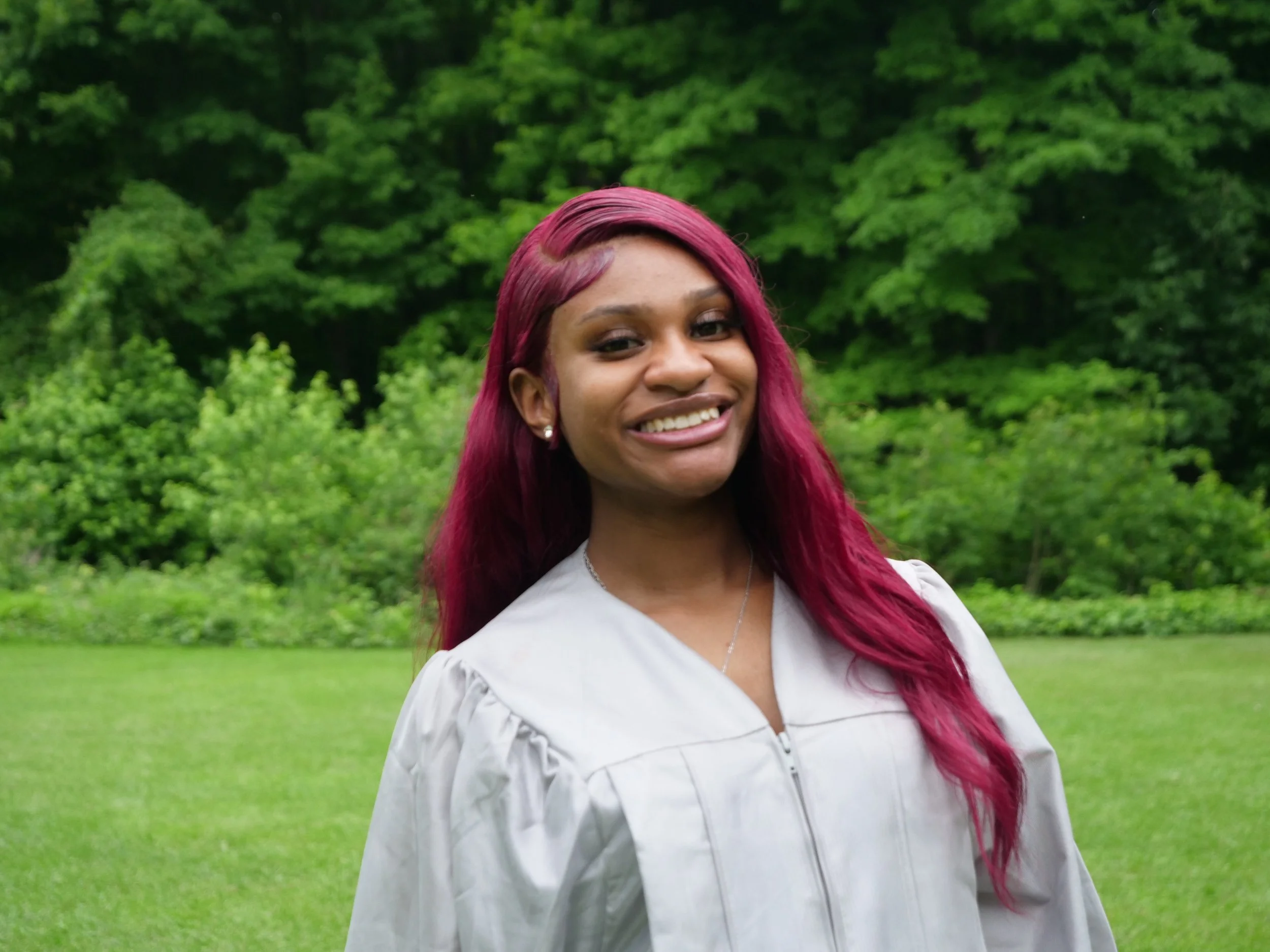 Young woman with long, wavy red hair smiling outdoors in front of green trees and grass, wearing a white graduation gown and jewelry.