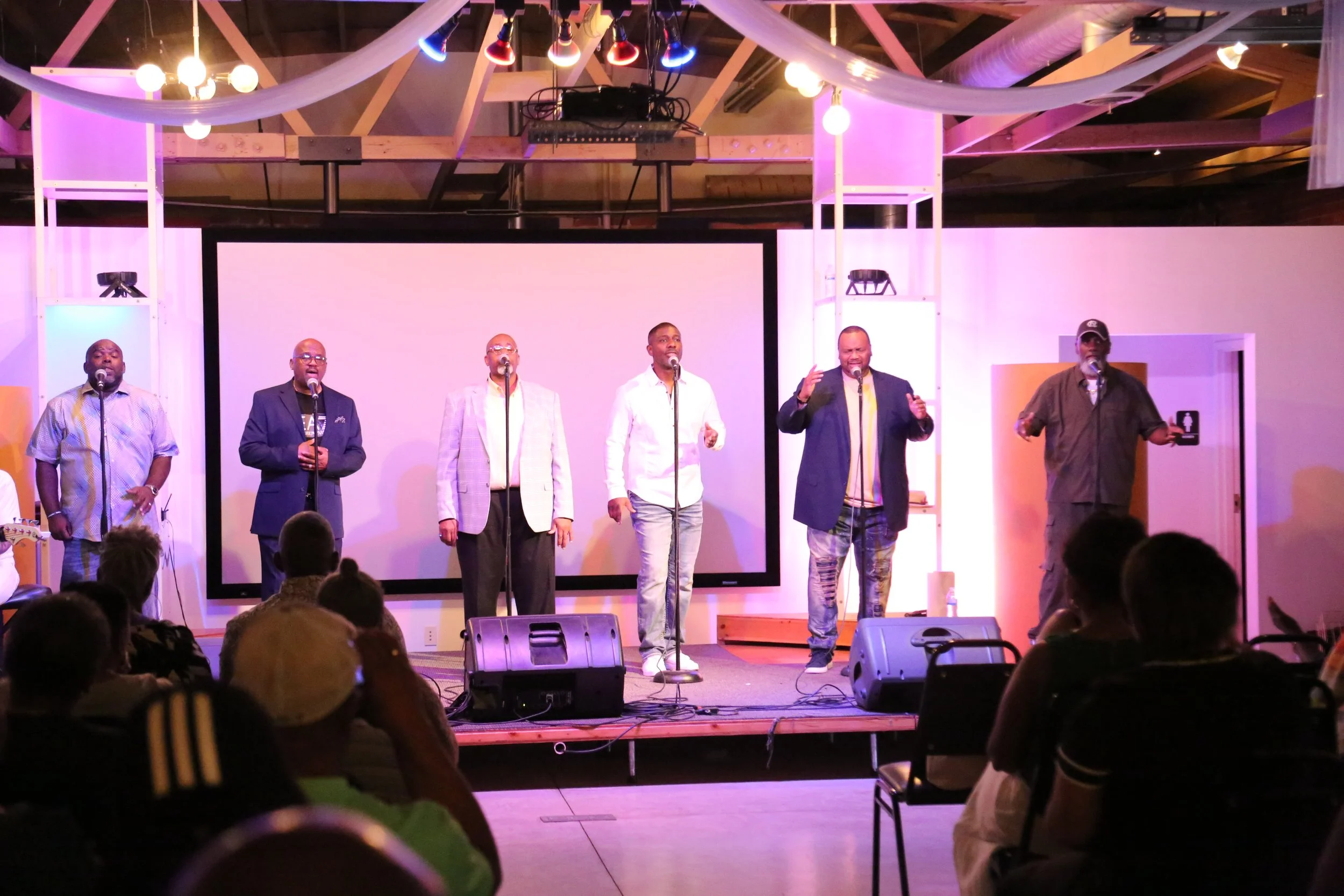 Six men singing on stage with a large blank screen behind them, stage lights overhead, and people in the audience watching.