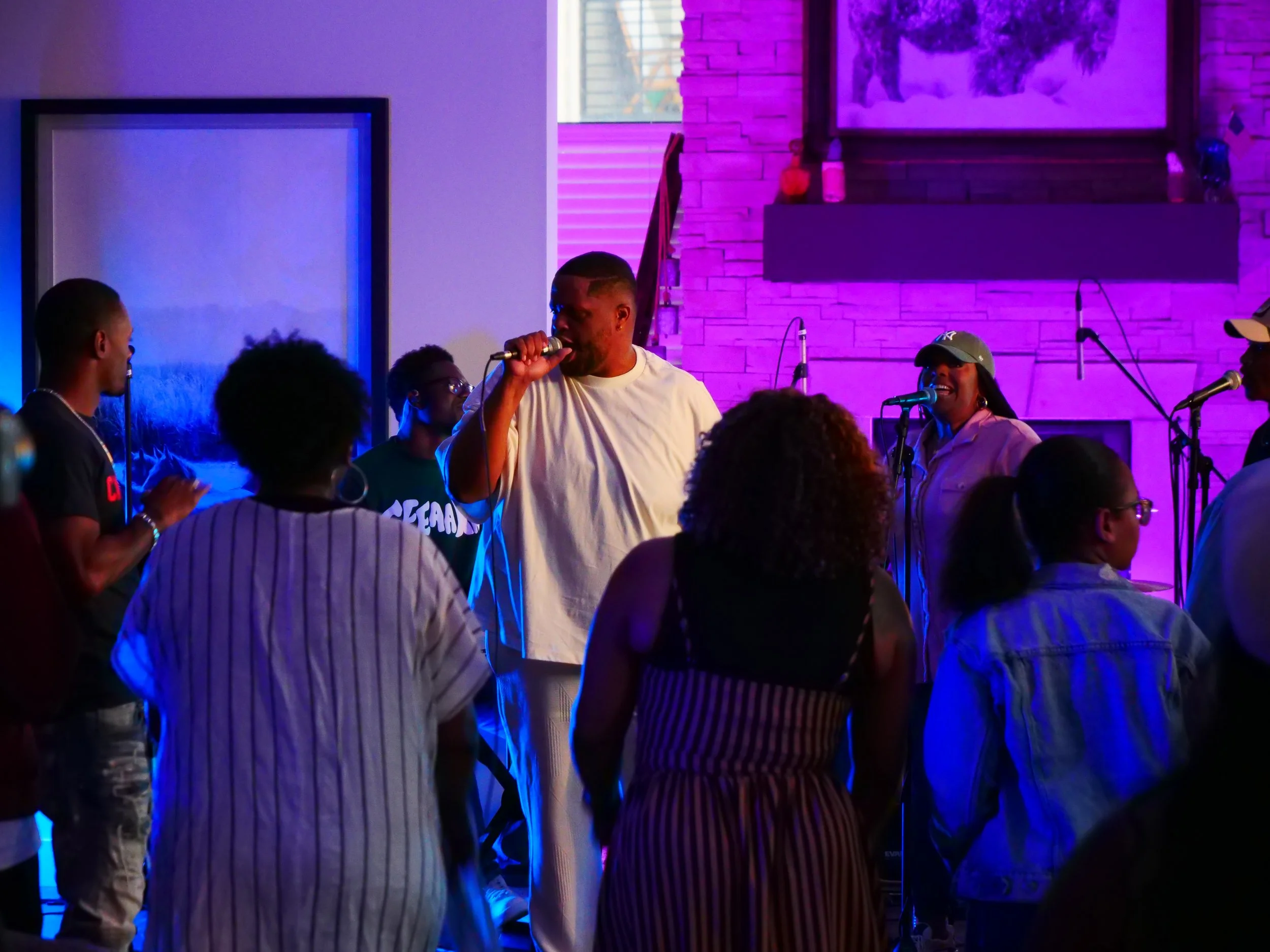 Group of people singing and dancing at a live music event in a cozy indoor venue with purple accent lighting, a brick fireplace, and scenic artwork on the wall.