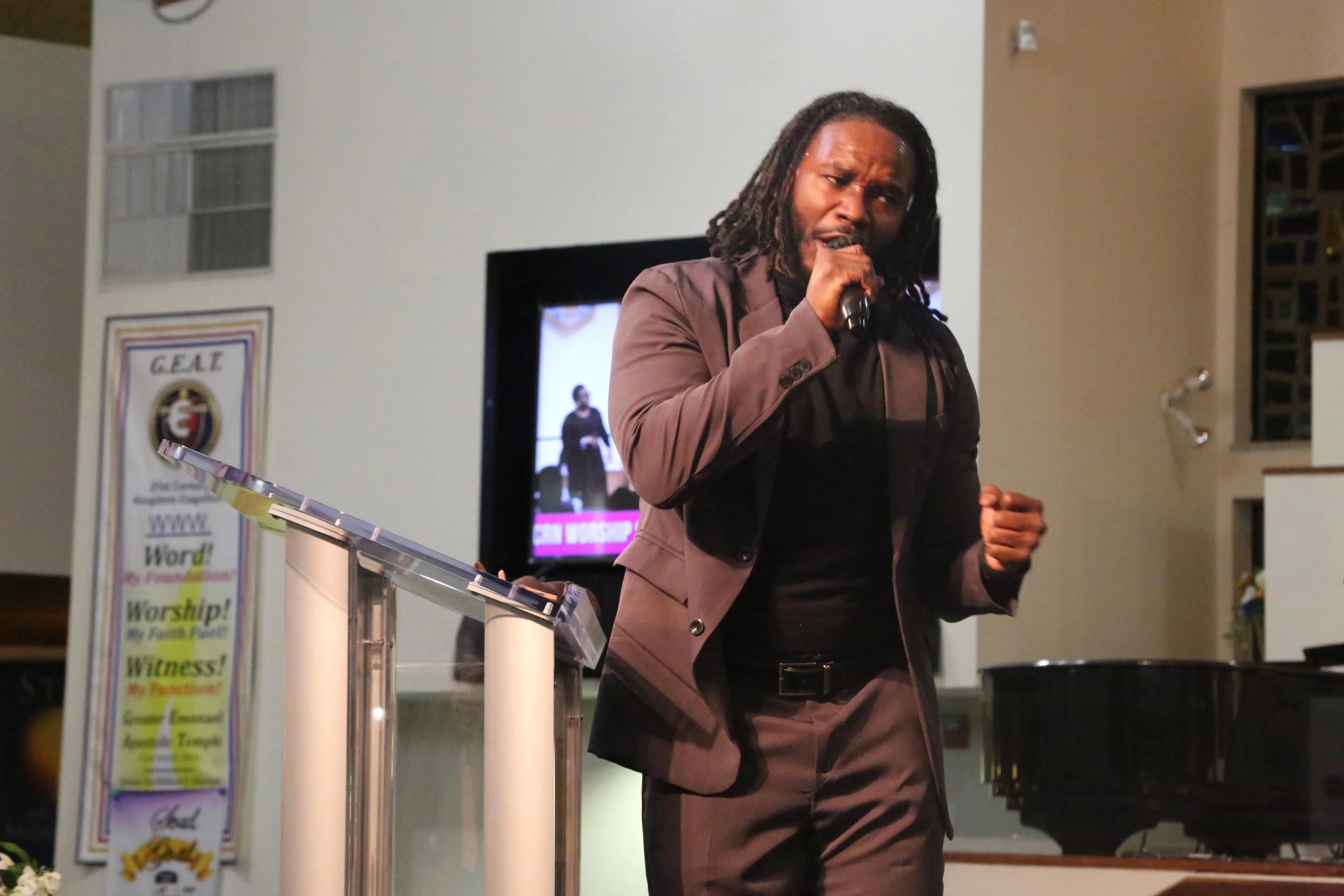 A man with dreadlocks singing into a microphone in a church or conference room, with a banner and television screen in the background.