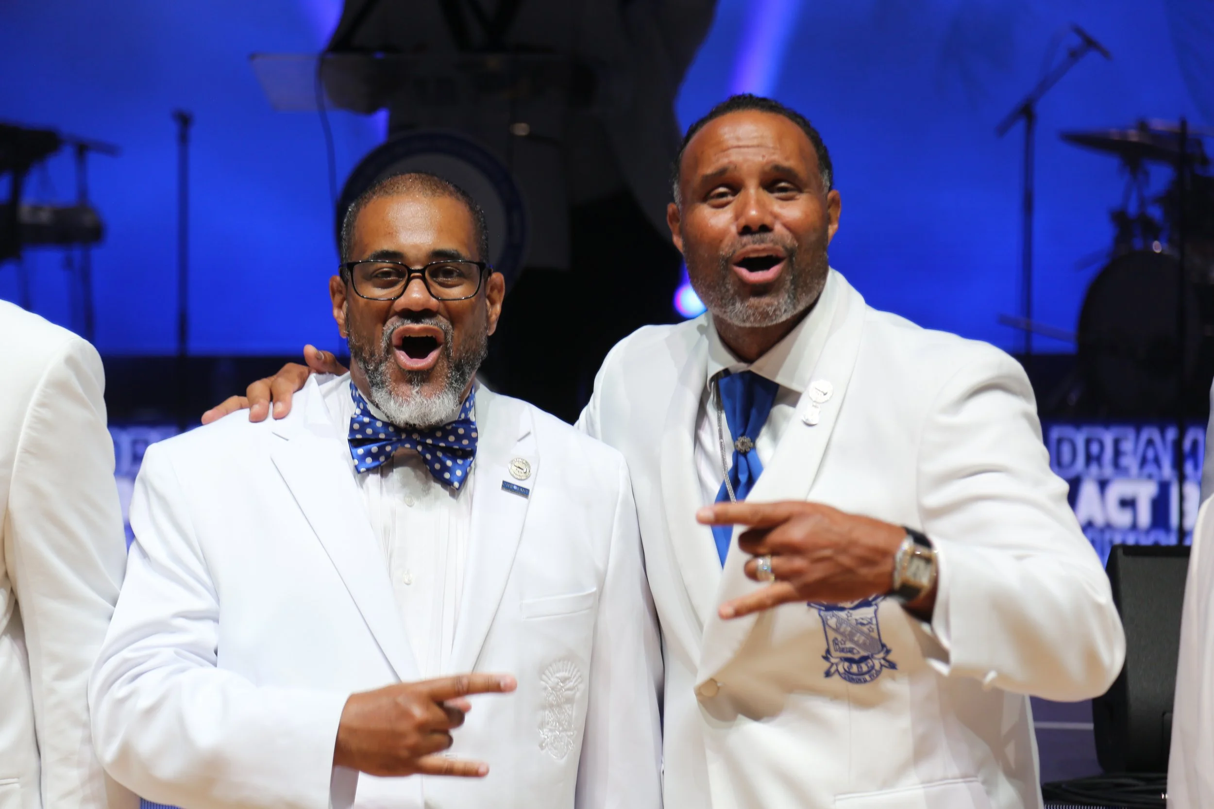 Two men in white suits with blue accessories celebrating at an event with a stage and musical instruments in the background.
