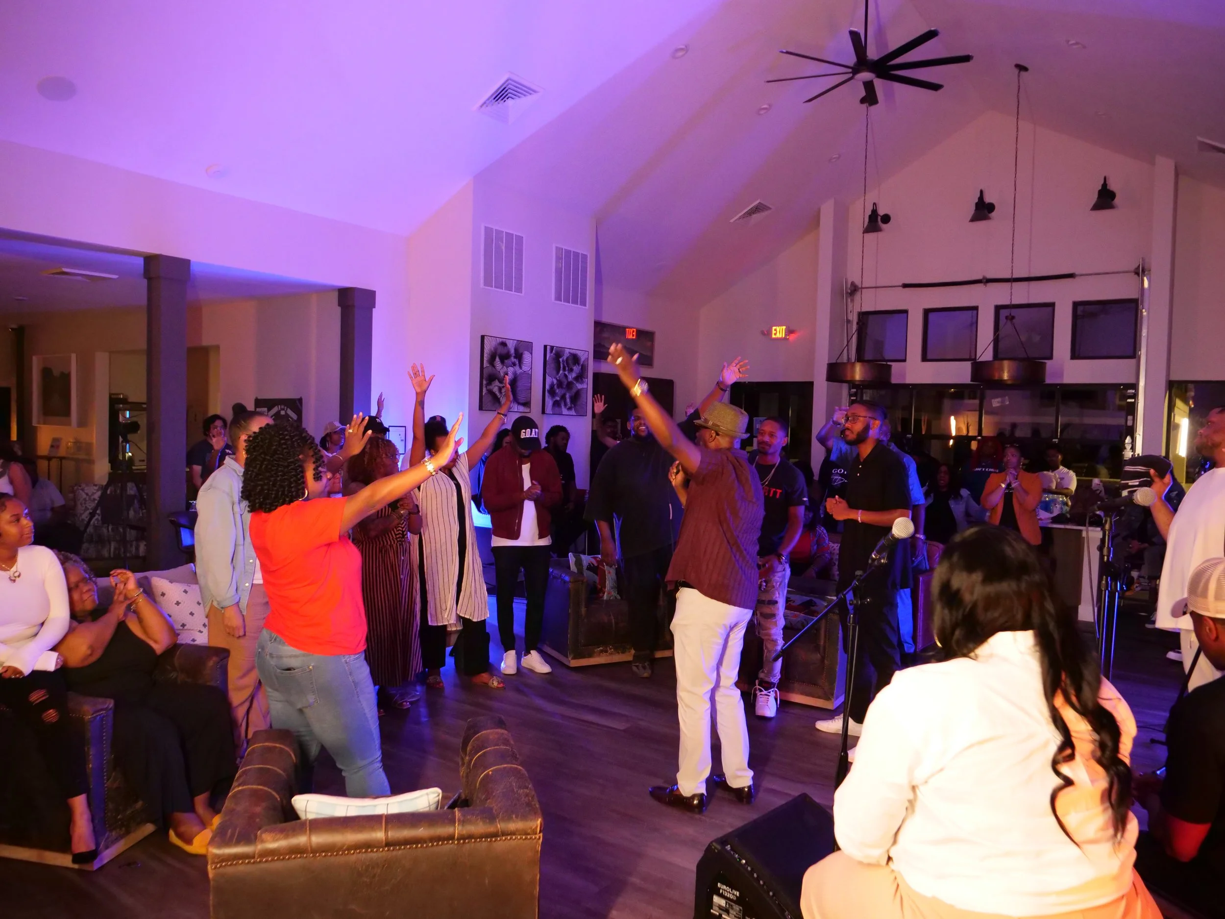 People dancing and socializing at an indoor event with purple lighting, some with hands raised, in a spacious room with modern decor and artwork on the walls.