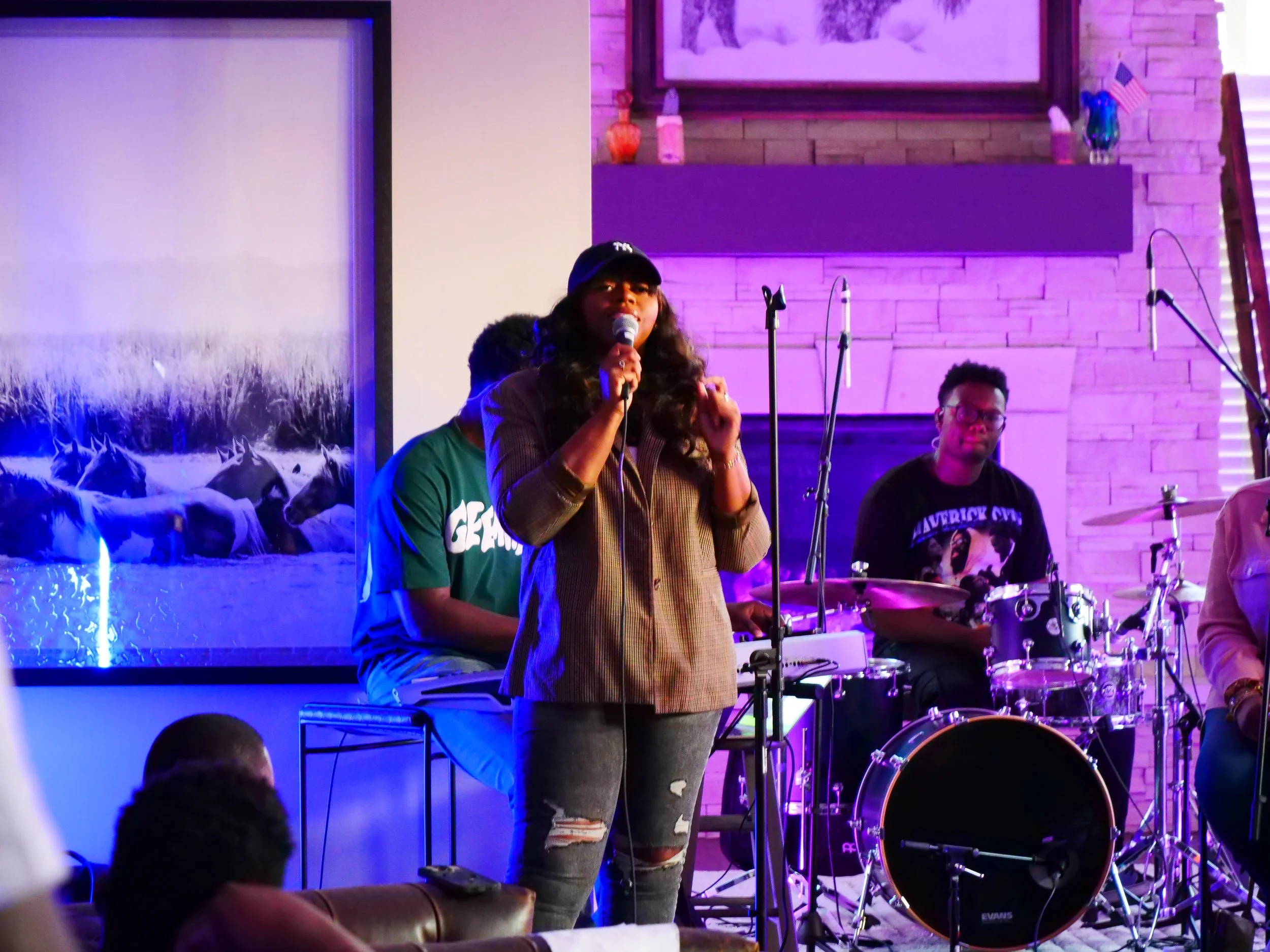 A woman with long dark hair, wearing a baseball cap and plaid shirt, holding a microphone and speaking on stage. Behind her are two musicians, one playing drums and the other on keyboard, in a room with purple lighting and a brick wall backdrop.