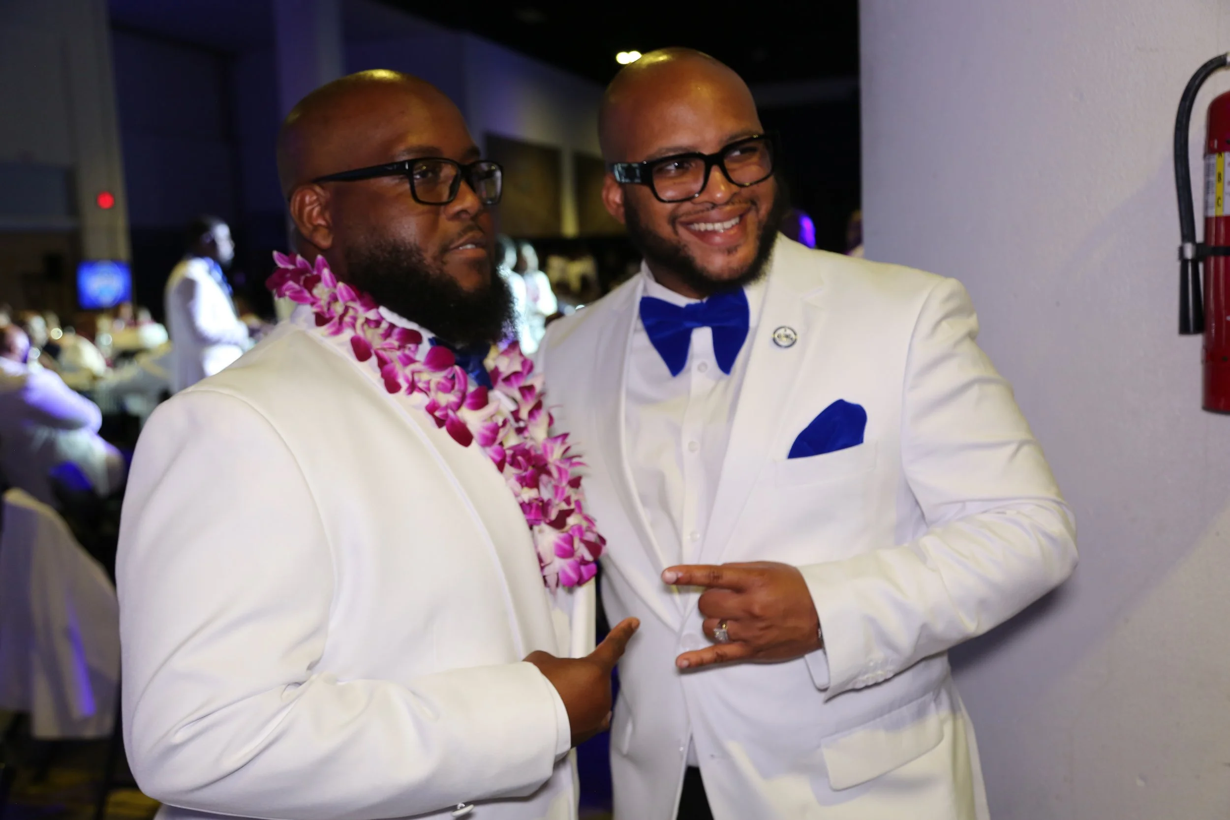 Two men dressed in white tuxedos with blue bow ties and glasses are at an indoor celebration. One man is wearing a purple lei and the other is pointing at him, smiling. There are other people in the background.