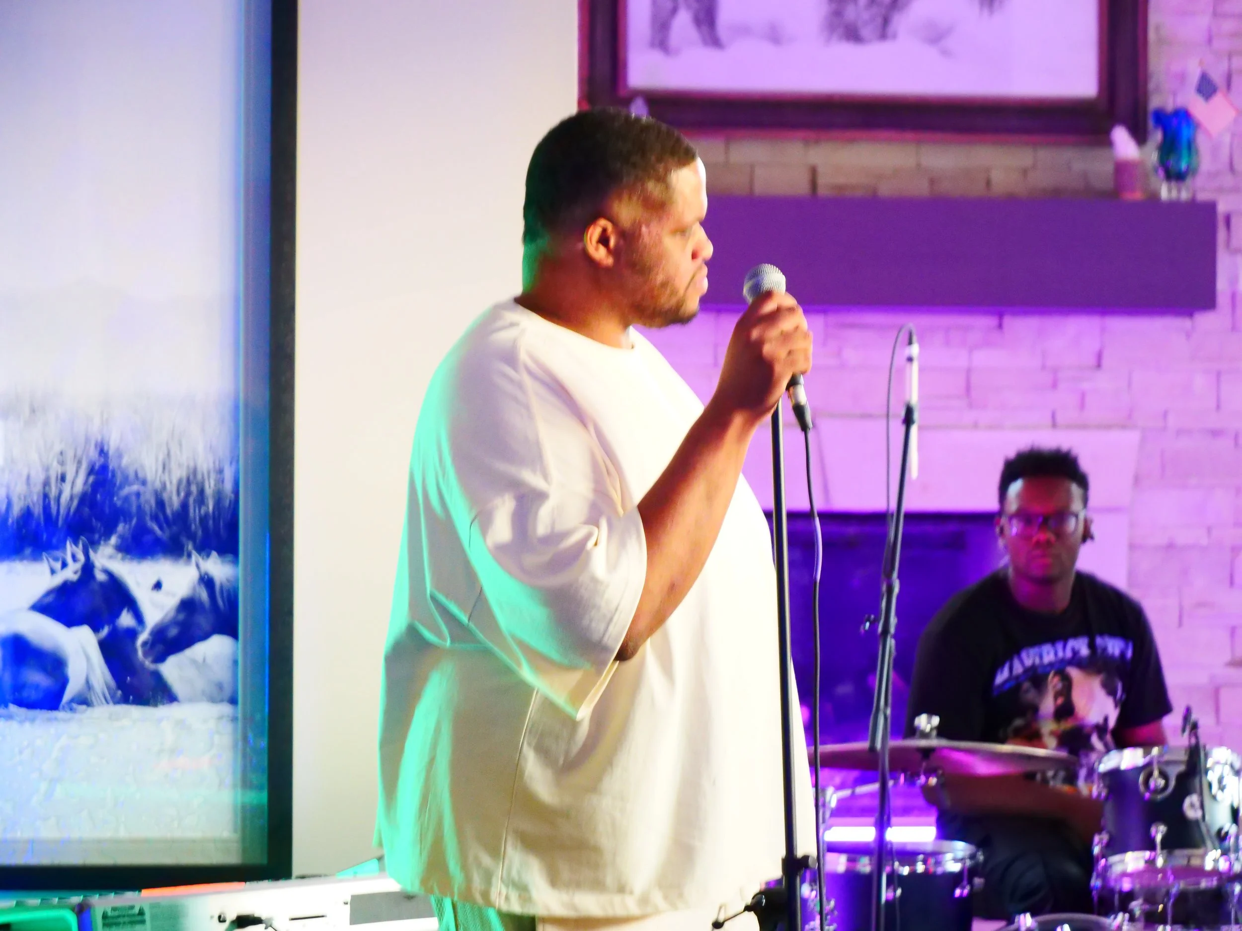 A man in a white shirt holding a microphone, singing or speaking, with a drummer in the background playing drums, in a room with purple and blue lighting and a brick wall.