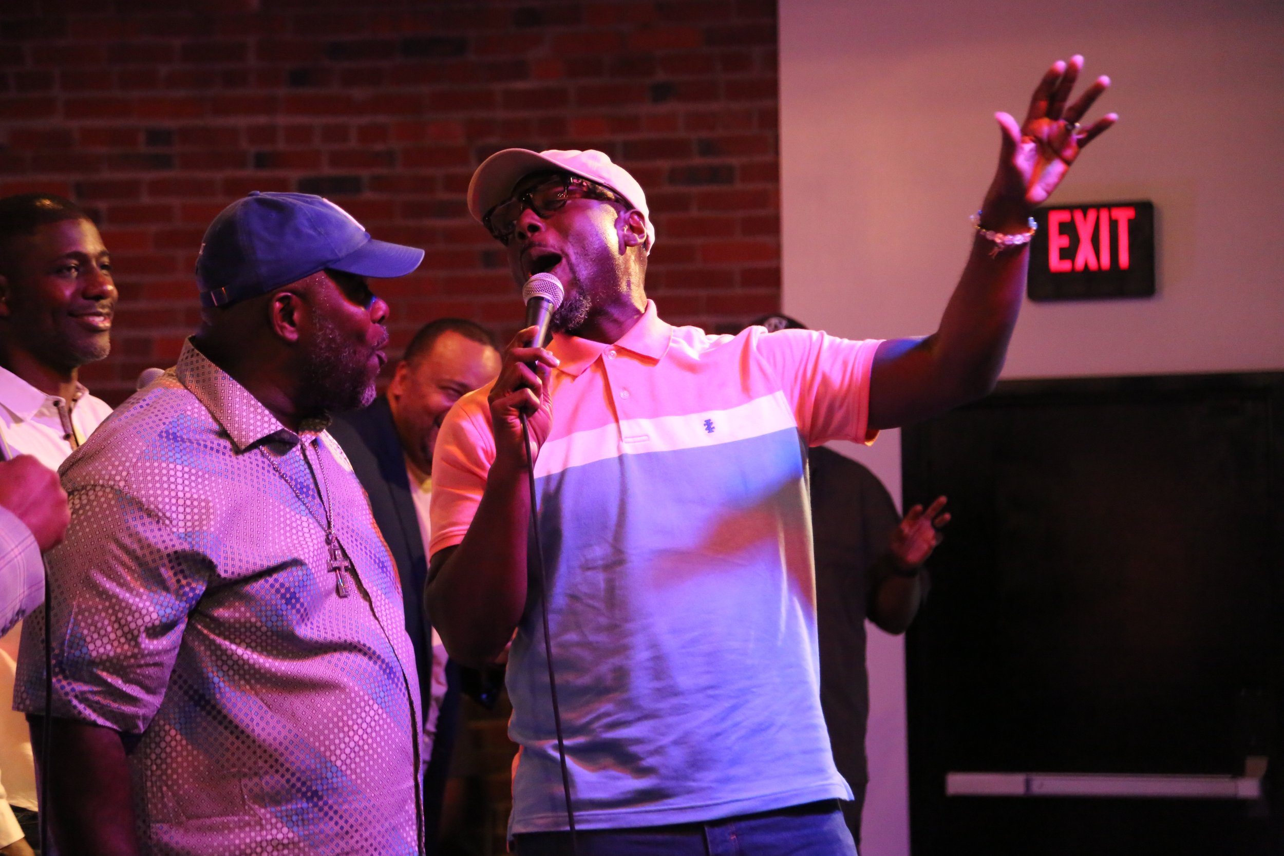 A man wearing a pink and blue polo shirt and a white cap is singing into a microphone with his right hand raised. Several other men are standing around him, smiling, against a brick wall background with an illuminated red exit sign.
