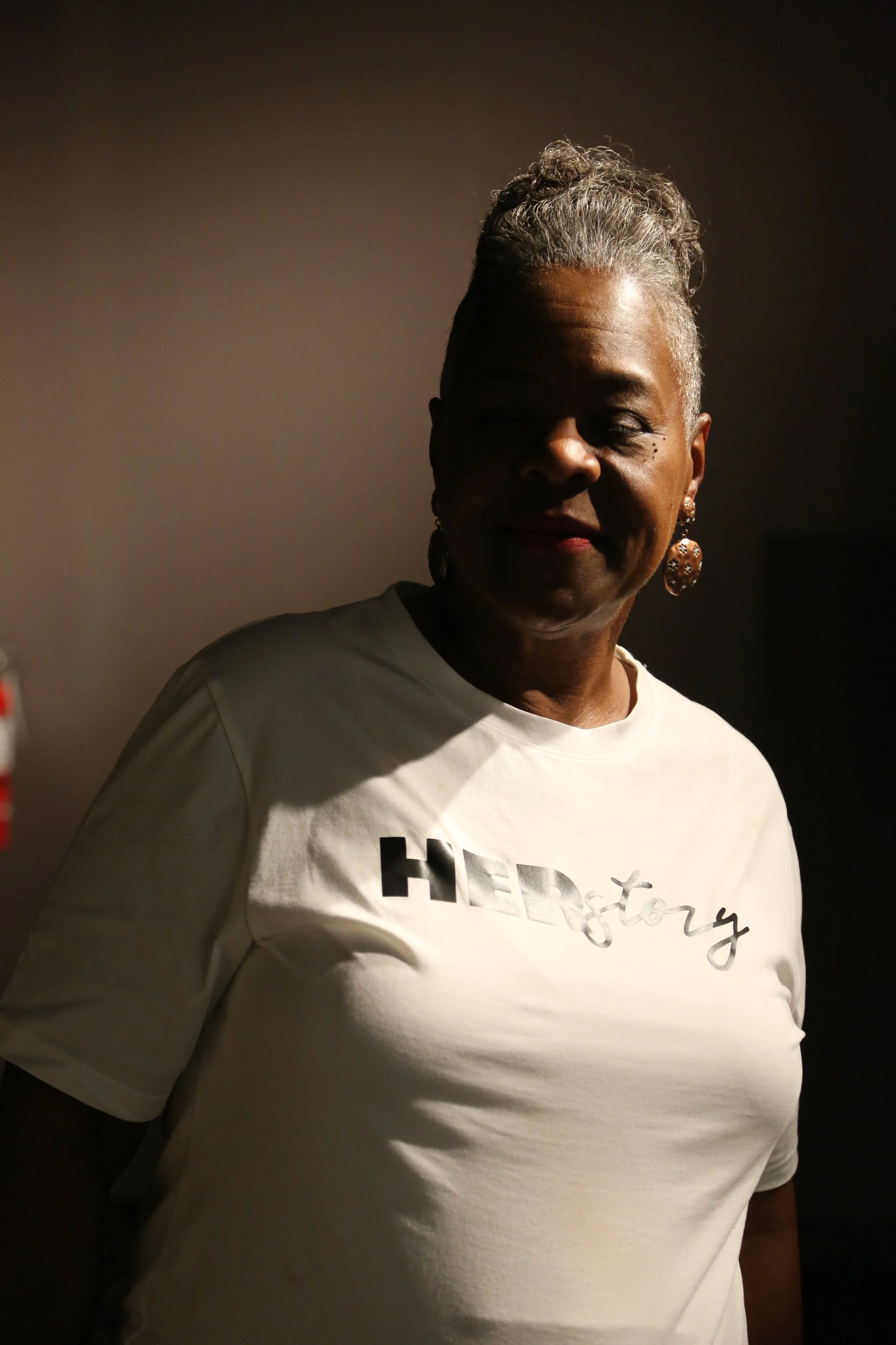 A woman with gray hair styled in an updo, wearing a white T-shirt with the word "HISTORY" printed on it, standing in a dimly lit room with a shadowed background.