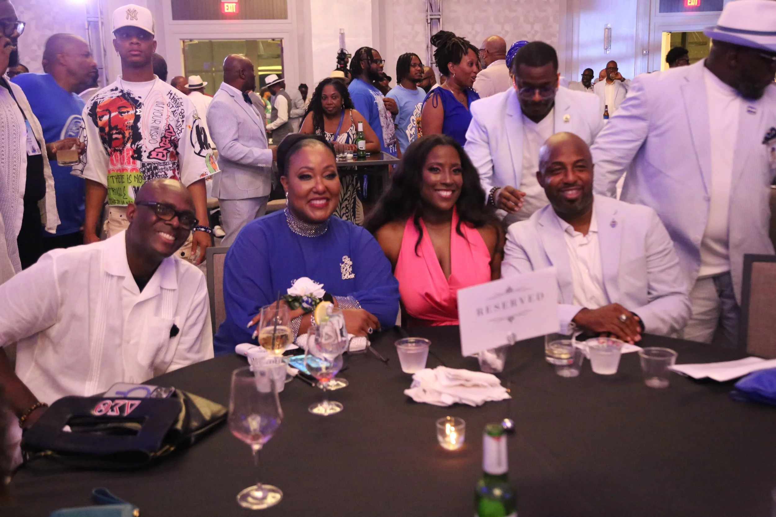 A group of people seated at a dinner table at a formal event, with some standing in the background. The table has drinks, a 'Reserved' sign, and a small candle.