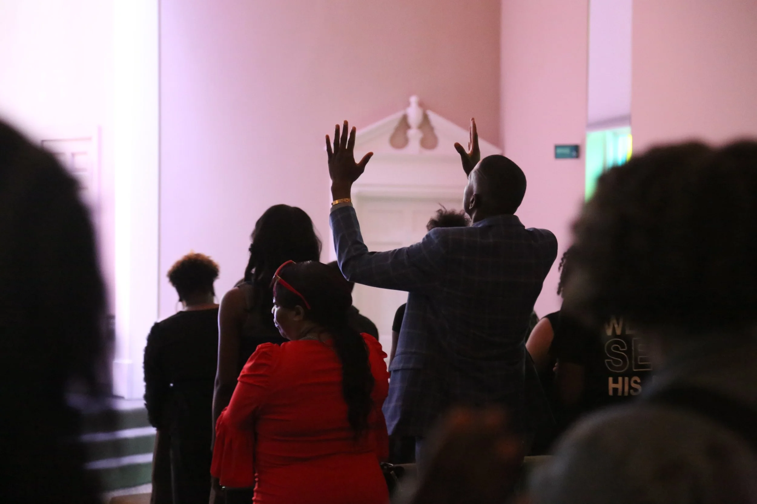 People standing in a hall with pink walls, some raising their hands, attending an event or gathering.