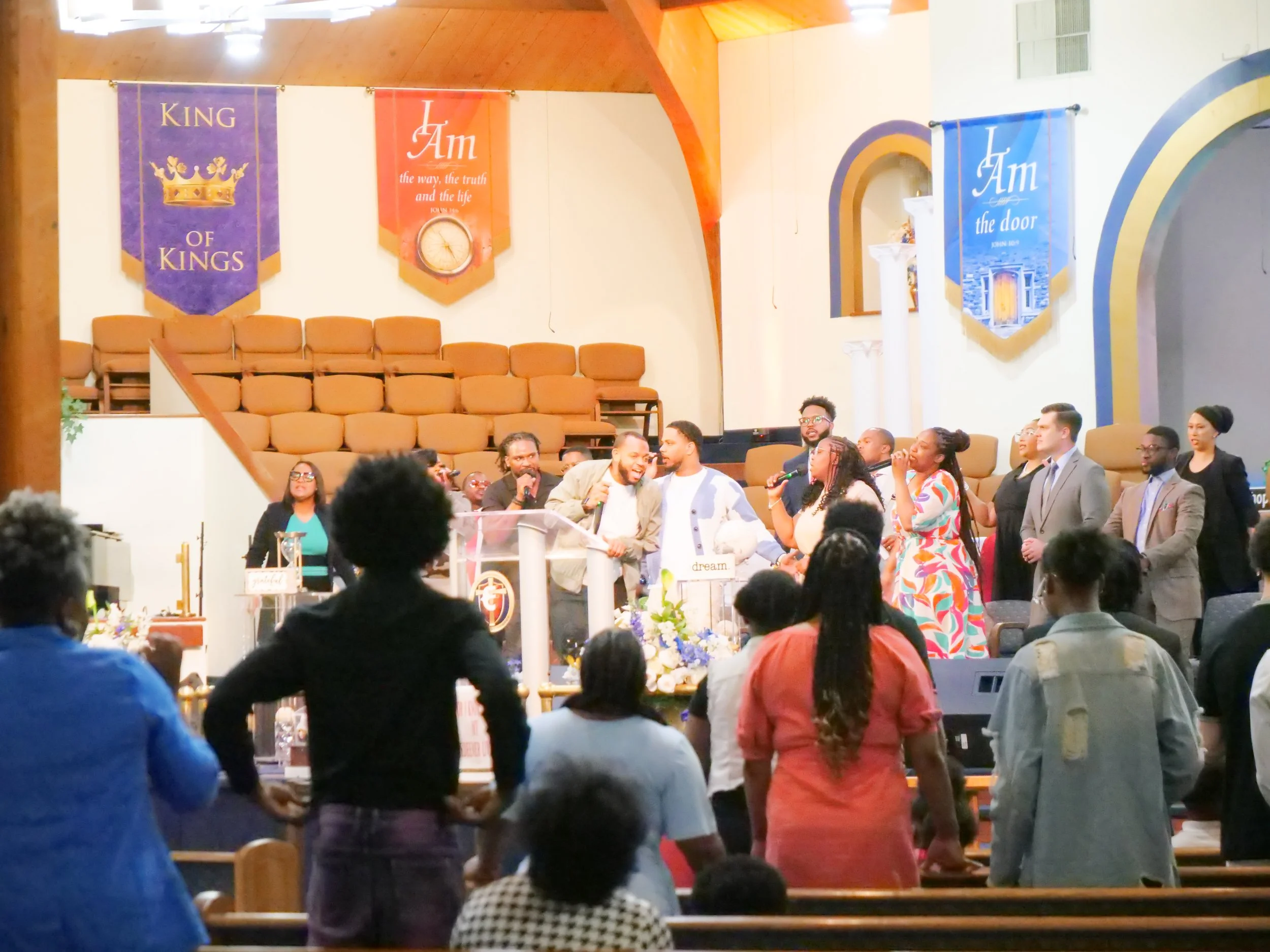 A diverse group of people gathered inside a church, standing at the front near the altar, with some singing and others listening. The church has banners on the wall that read 'King of Kings,' 'I Am,' and 'I Am the door.'