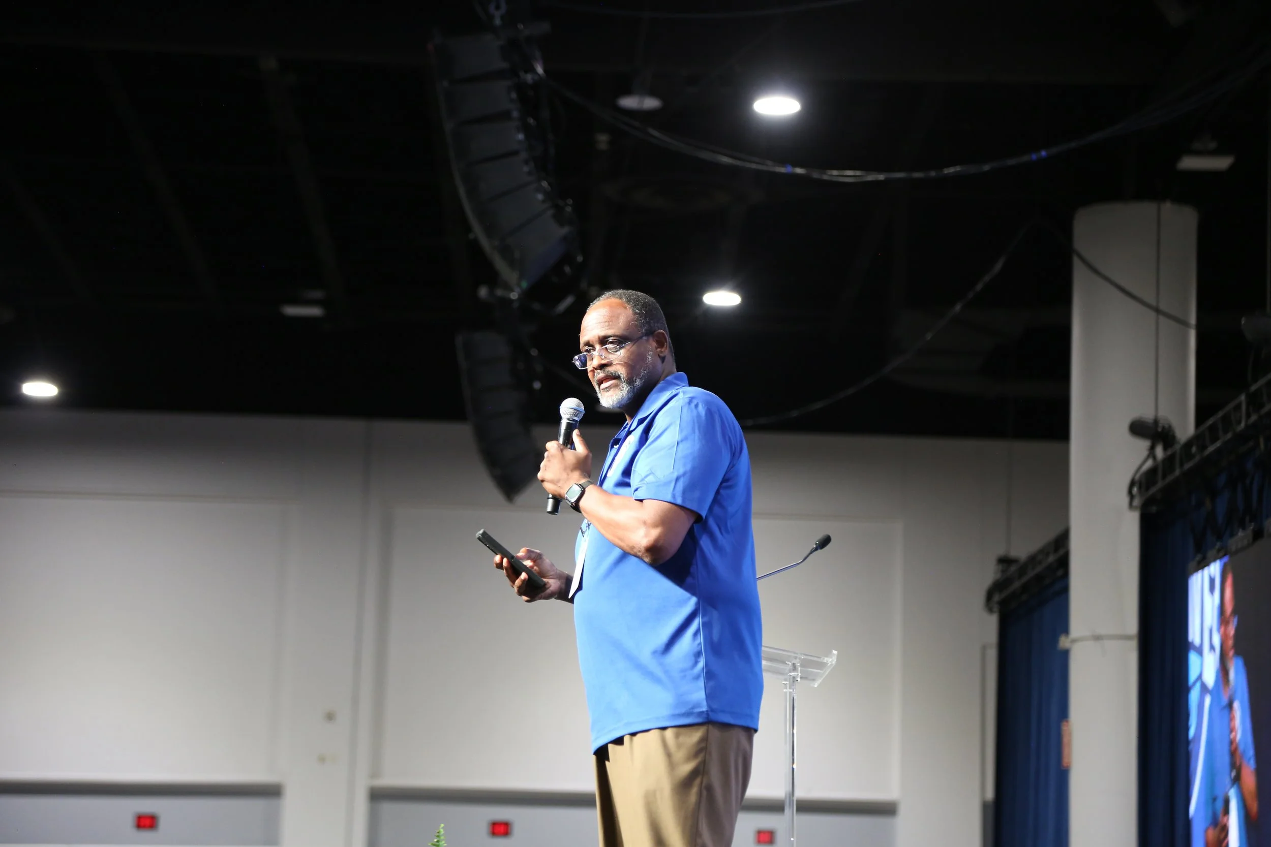 A man in blue shirt and khaki pants standing on stage, speaking into a microphone, holding a smartphone, with a transparent podium in front, and large speakers hanging above.