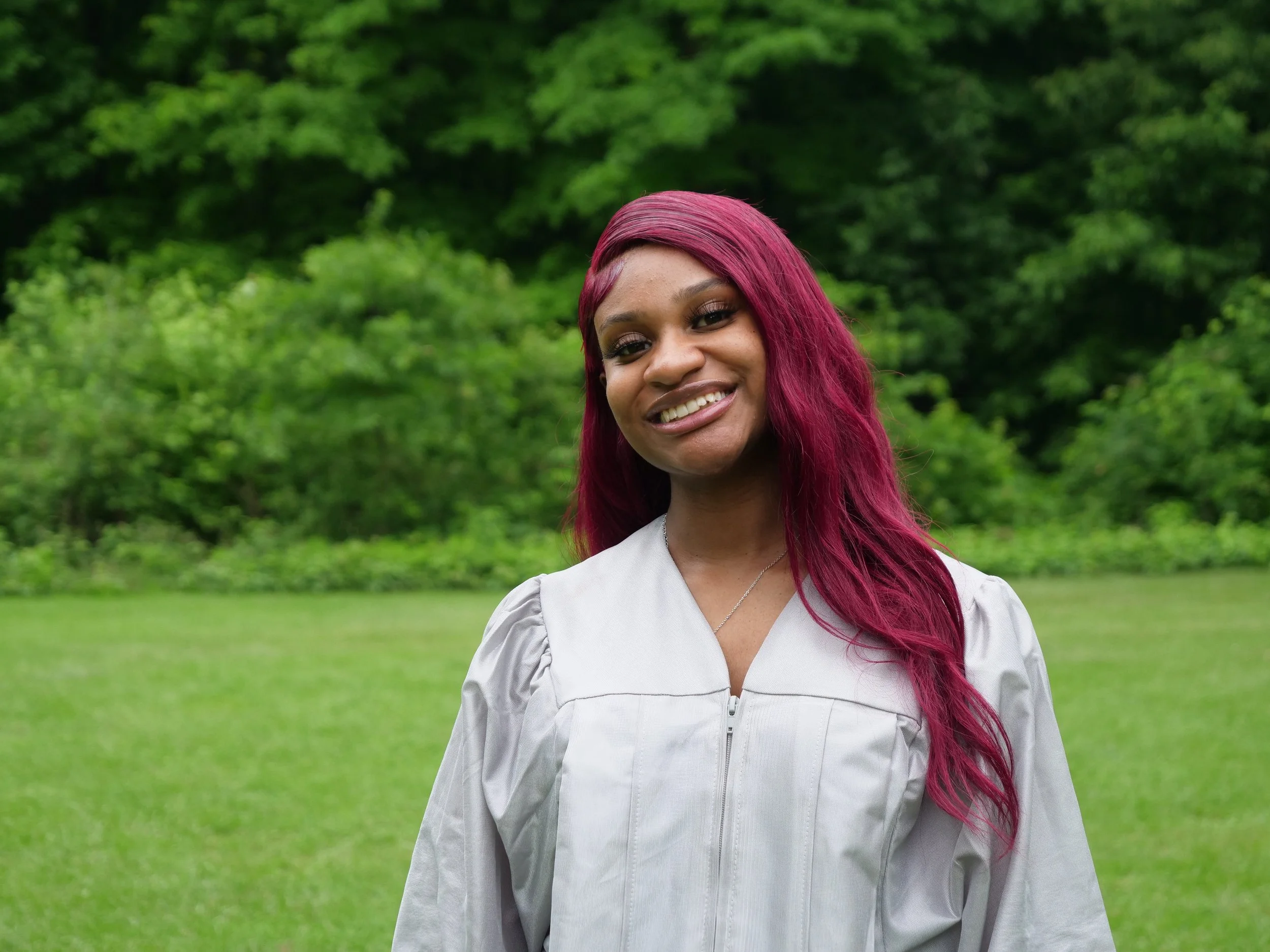 Young woman with long, wavy burgundy hair smiling in a lush green park.