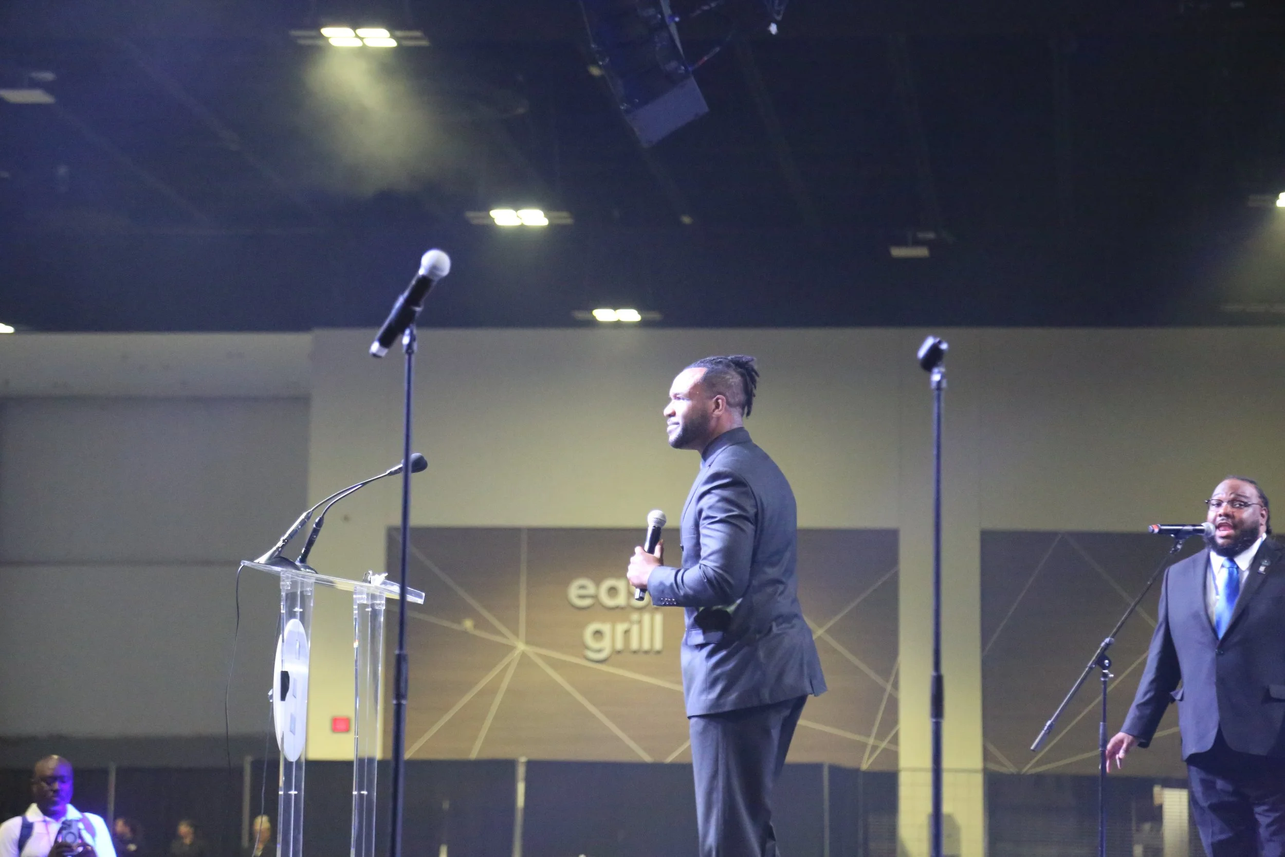 Two men in suits standing on stage holding microphones, with one man smiling and facing sideways, and another man also holding a microphone, in front of a sign that reads 'eat grill' on a plain yellow and brown background, with audience members in th