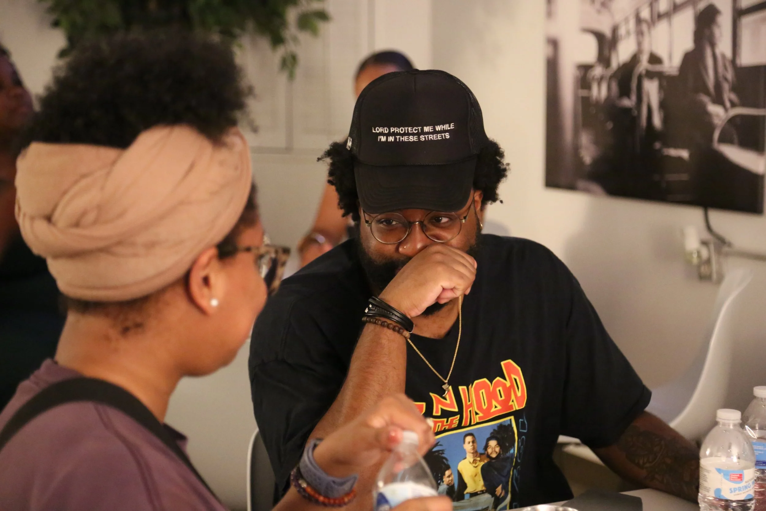 Two people sitting at a table in a casual indoor setting having a conversation. One woman with a tan headwrap and glasses, and a man with black baseball cap, glasses, a Black T-shirt, and bracelets.