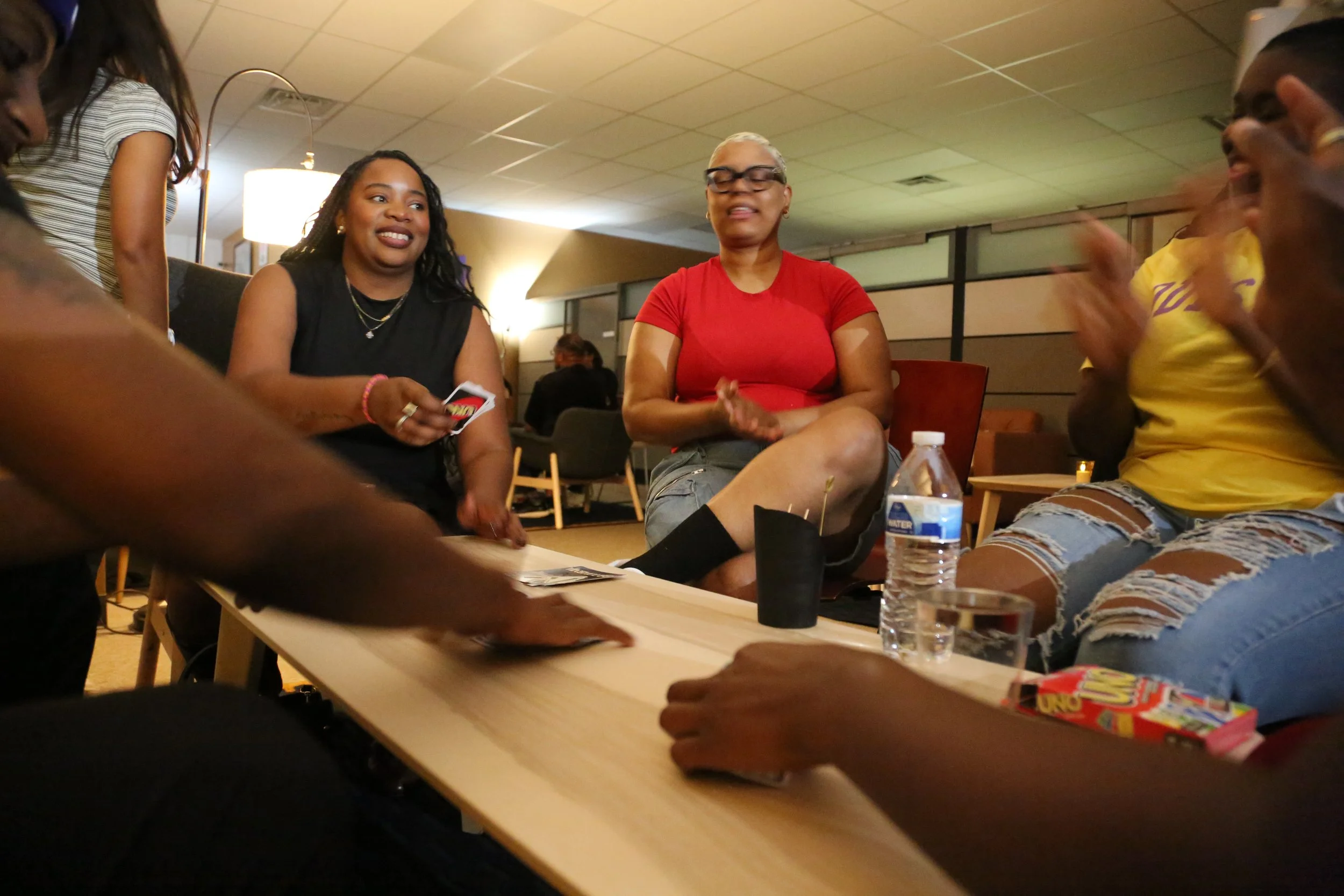Group of people sitting around a table, smiling, playing a card game, with a water bottle and UNO game on the table, in a cozy indoor setting.