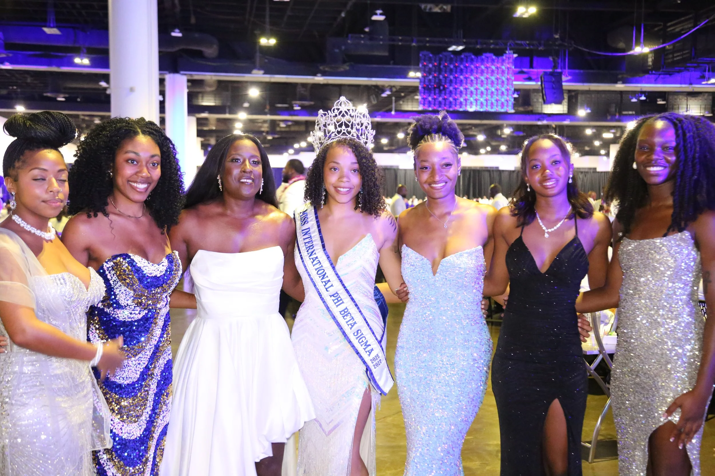 A group of seven women dressed in formal evening gowns standing together at an indoor event, including a woman with a crown and sash indicating she is Miss International Pet Belt Belize 2022. They are smiling and posing for the photo.