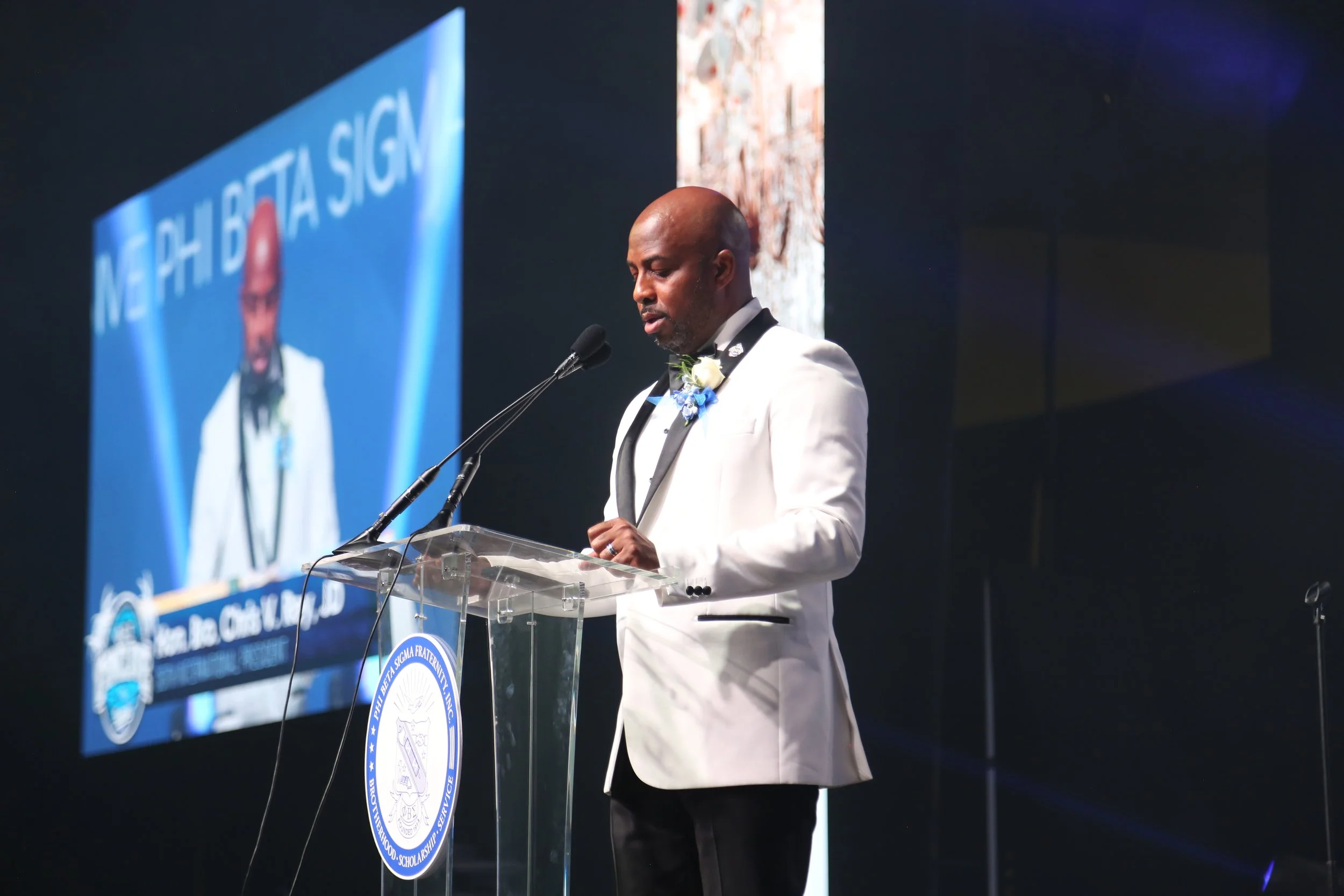 A man in a white tuxedo jacket giving a speech at a podium during a formal event. There is a large screen behind him displaying his image and the event's logo.