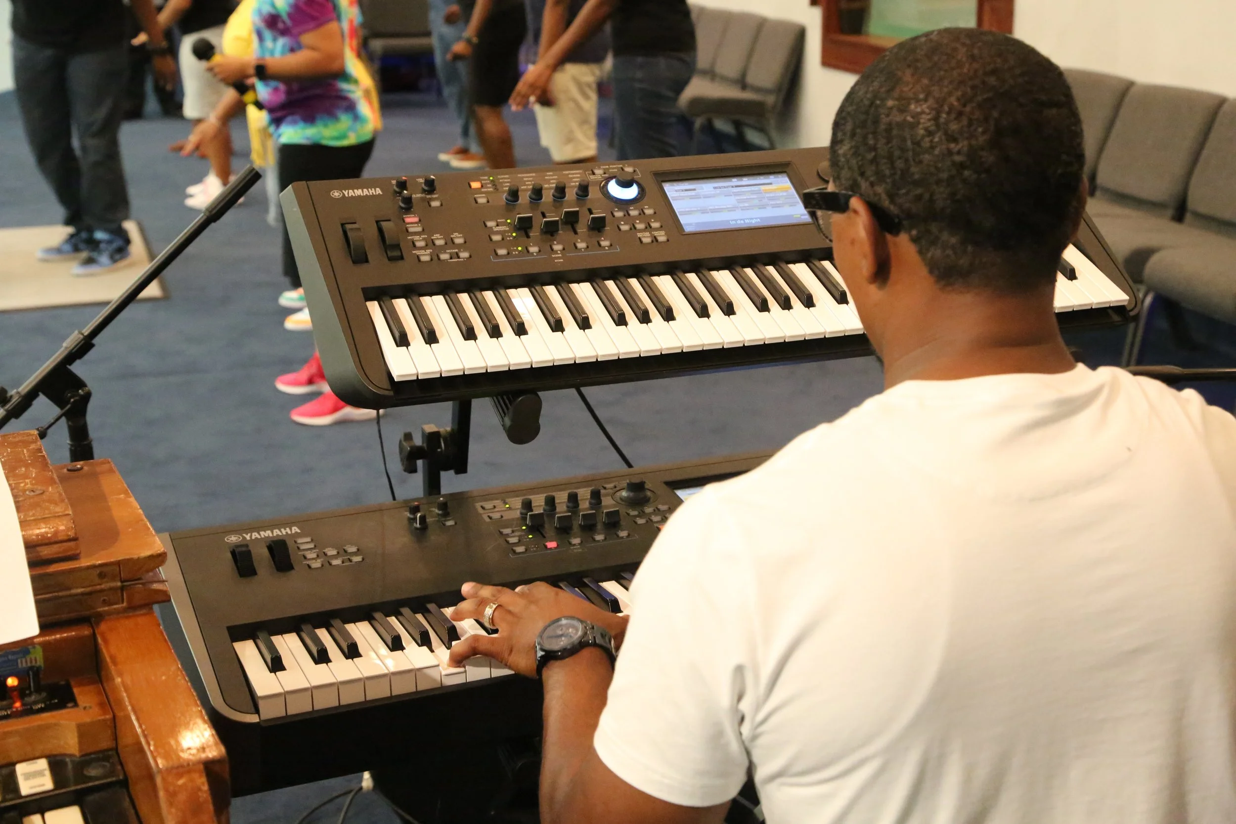 A man wearing a white t-shirt and glasses playing a Yamaha keyboard in a room with chairs and people in the background.