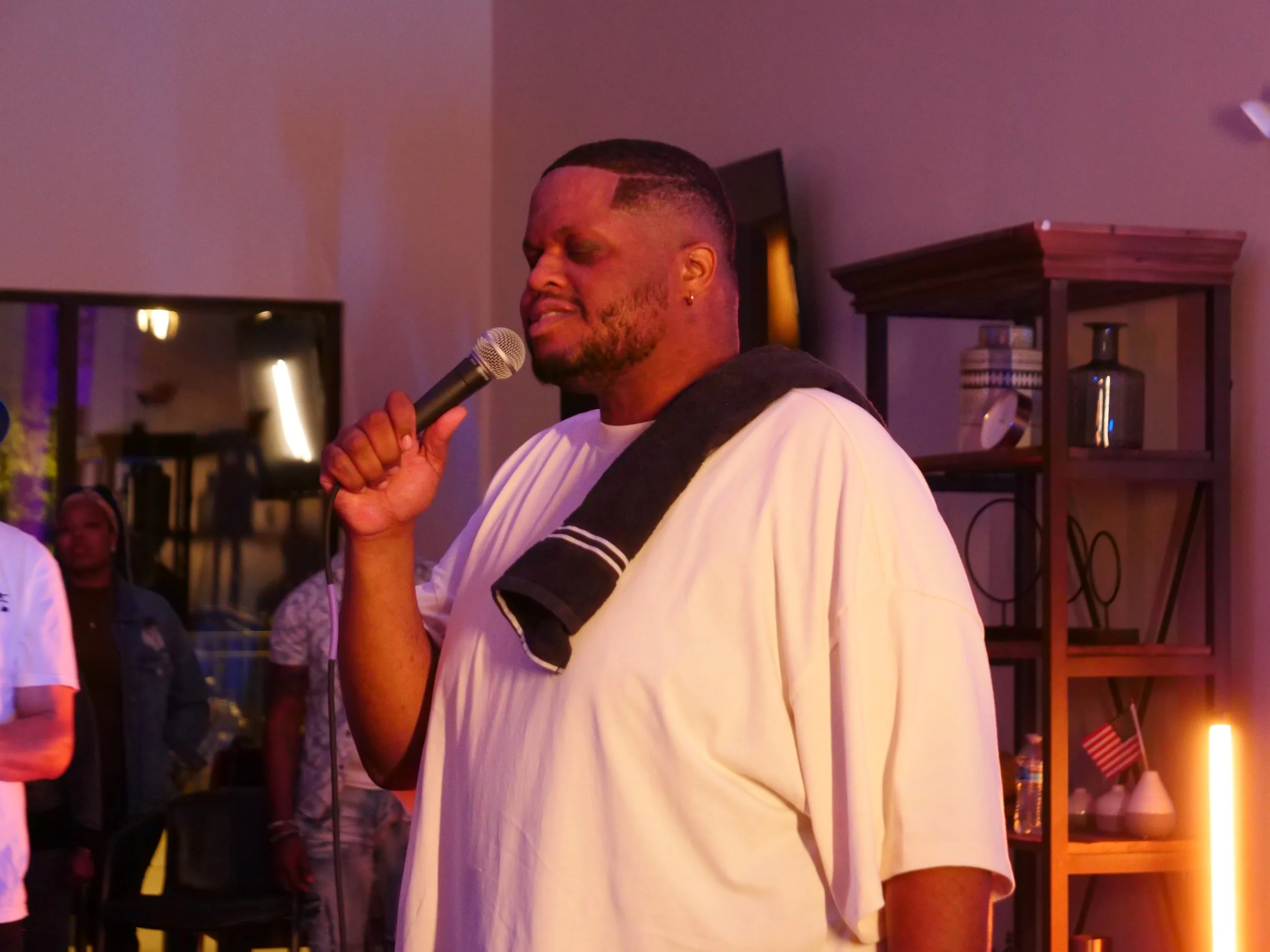A man with a beard and short hairstyle holding a microphone, with his eyes closed, appearing to sing or speak, wearing a light-colored t-shirt and a black sweatshirt draped over his shoulder in an indoor setting.