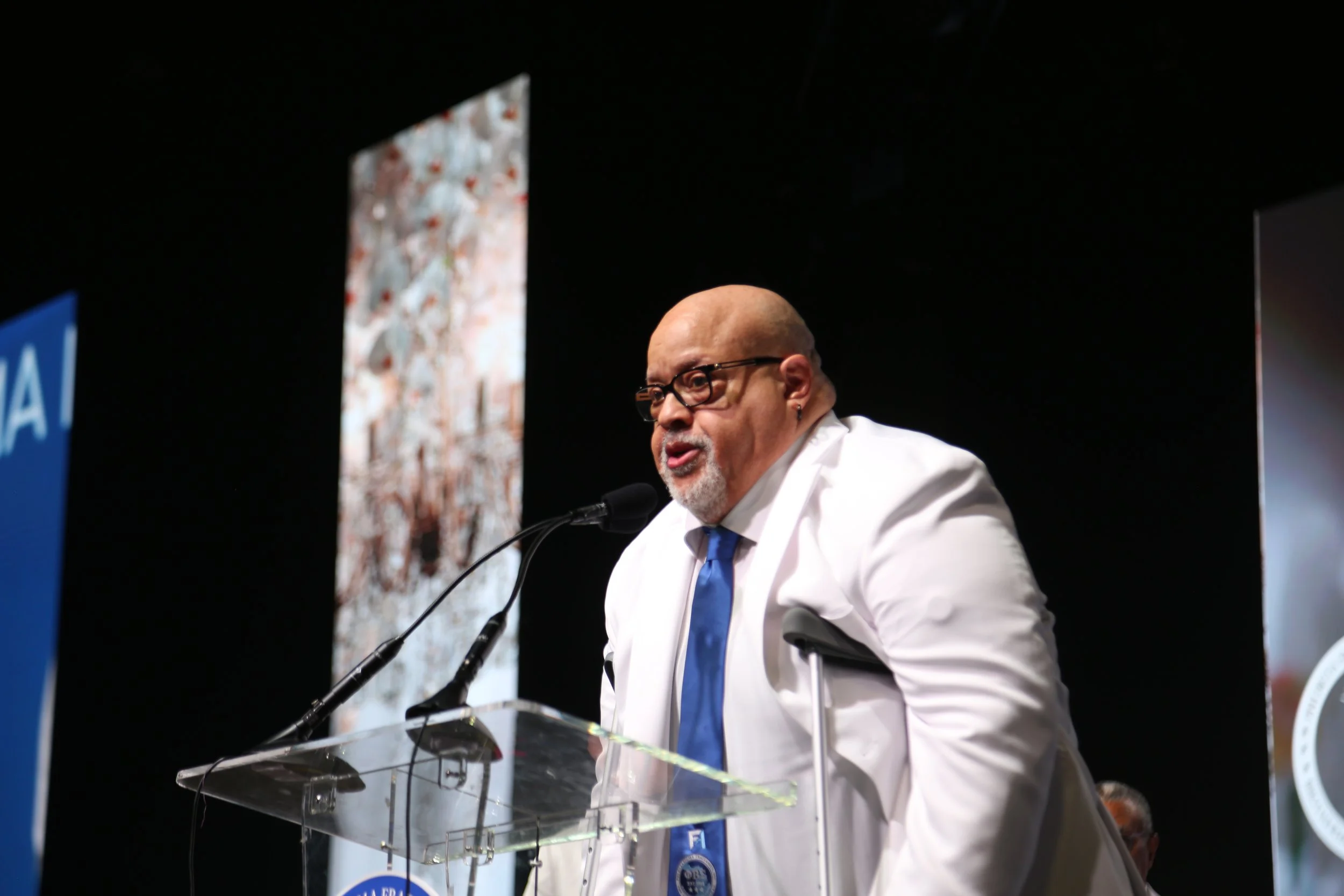 A man with glasses, a bald head, and a beard, dressed in a white suit and blue tie, speaking at a podium with microphones during a conference or event.
