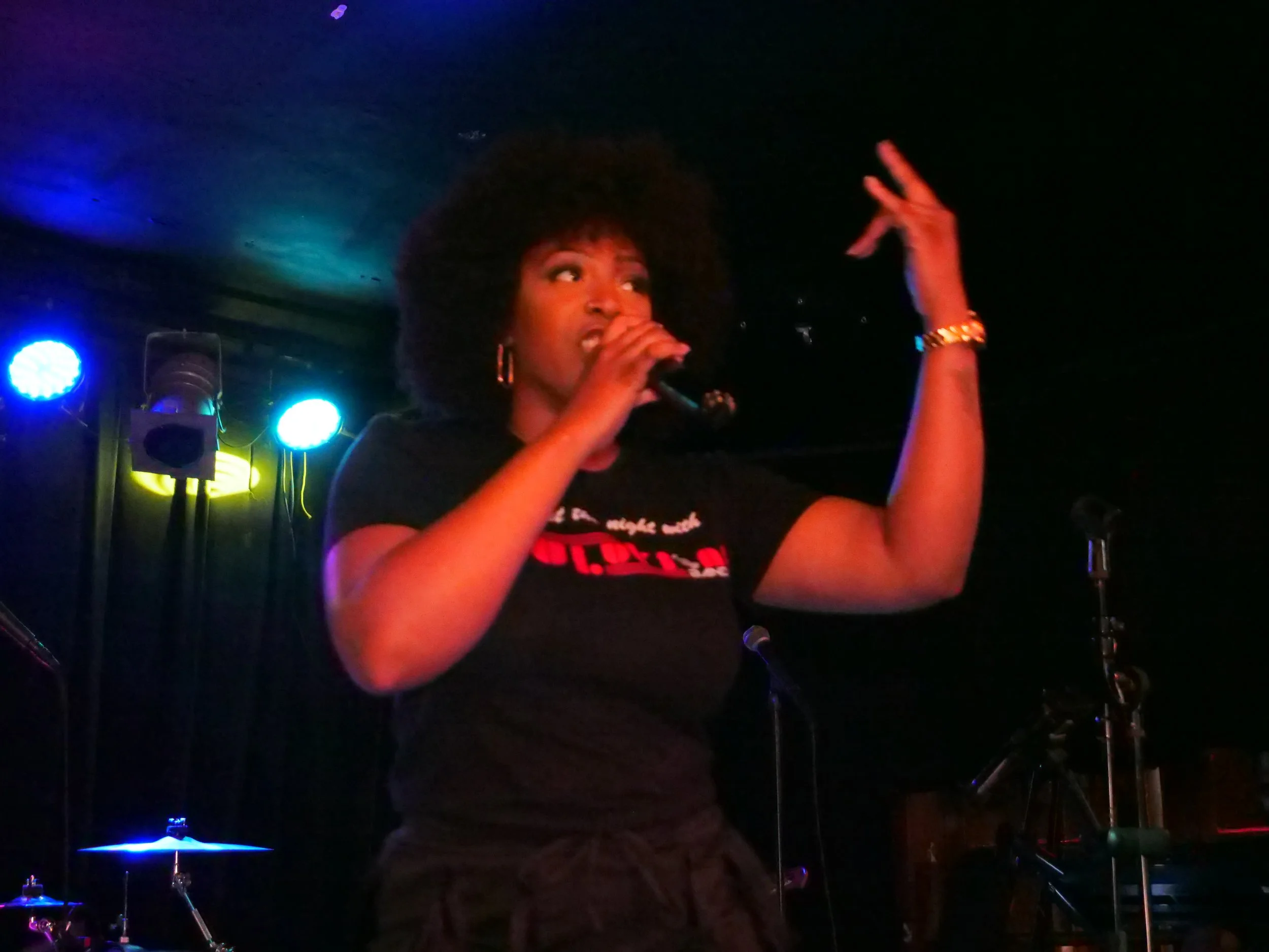 A woman with curly hair, wearing a black shirt and gold jewelry, standing on stage with a microphone in her hand, performing in a dark, colorful-lit venue.
