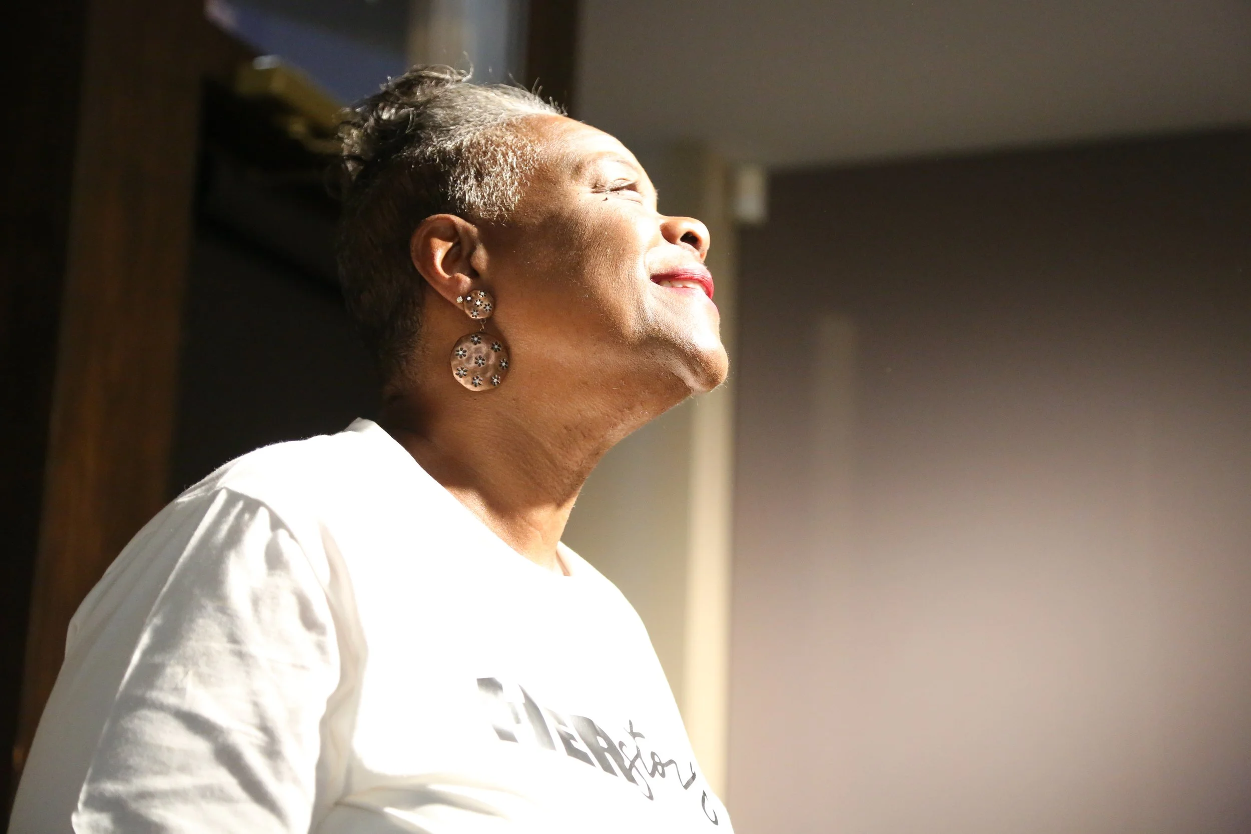 An older woman with short gray hair, wearing large earrings and a white shirt, is smiling with her eyes closed as sunlight shines on her face.