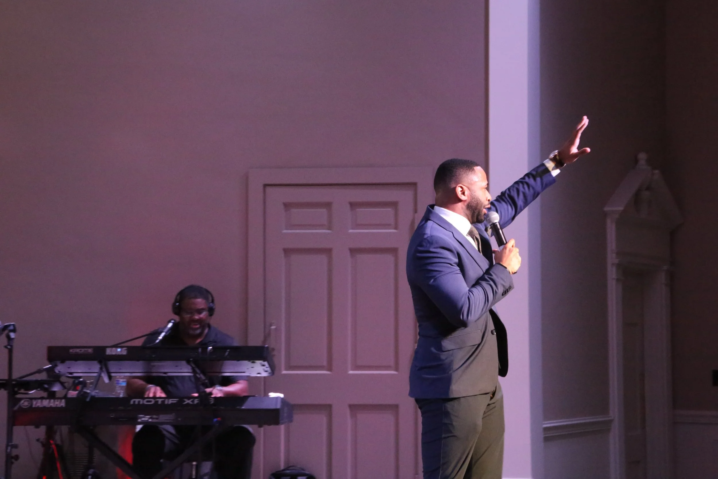 A man in a navy suit speaking into a microphone on stage, raising his right hand, while a keyboard player in the background performs and wears headphones.