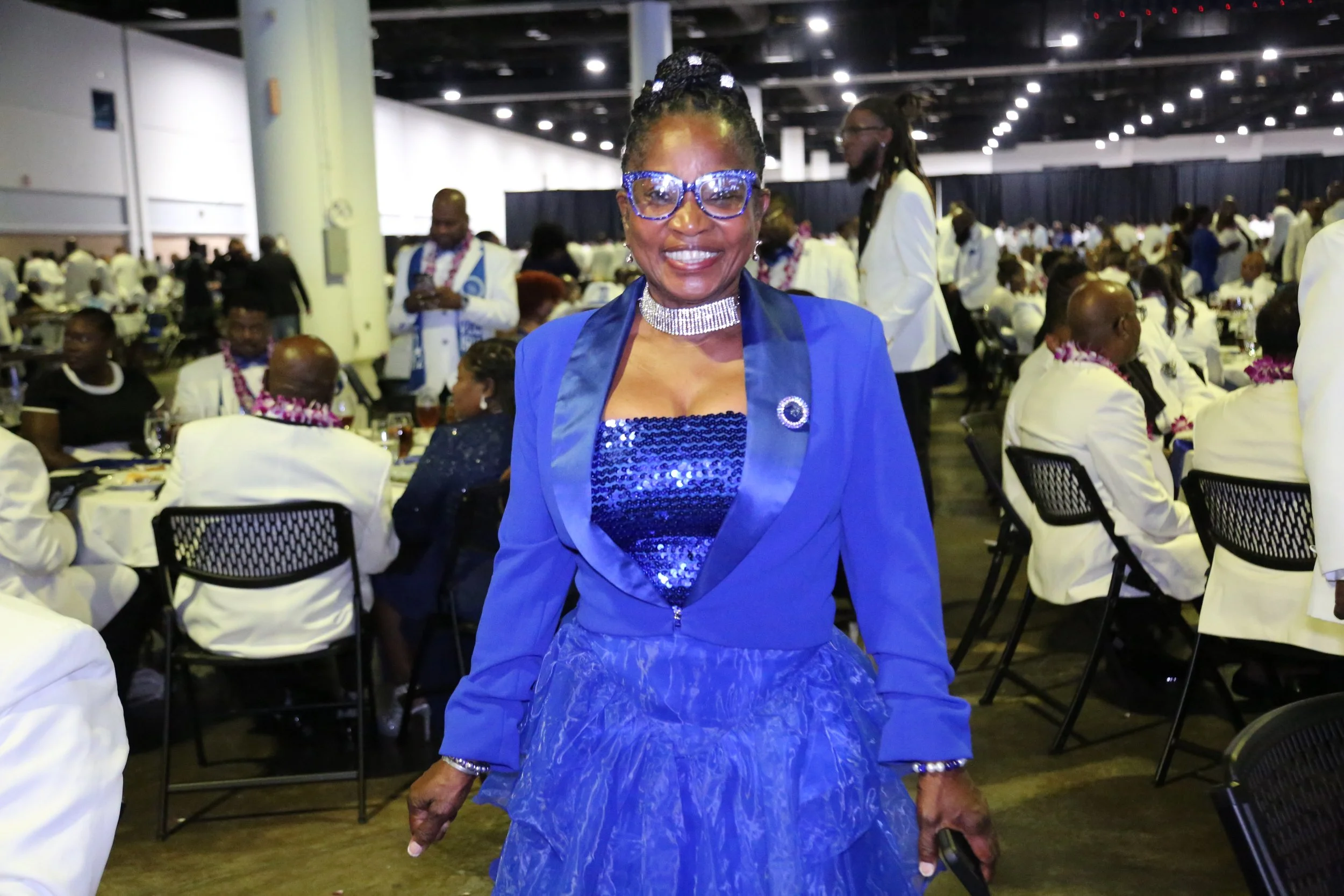 A smiling woman in a blue outfit and glasses standing at a formal event with many people seated at tables, dressed in white and black, in a large banquet hall.