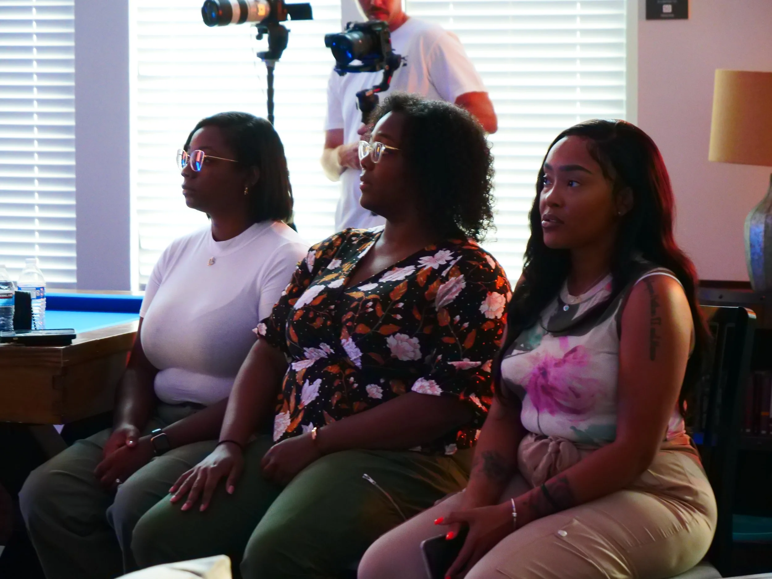 Three women sitting in a row during a presentation or event, with a man operating a camera in the background.