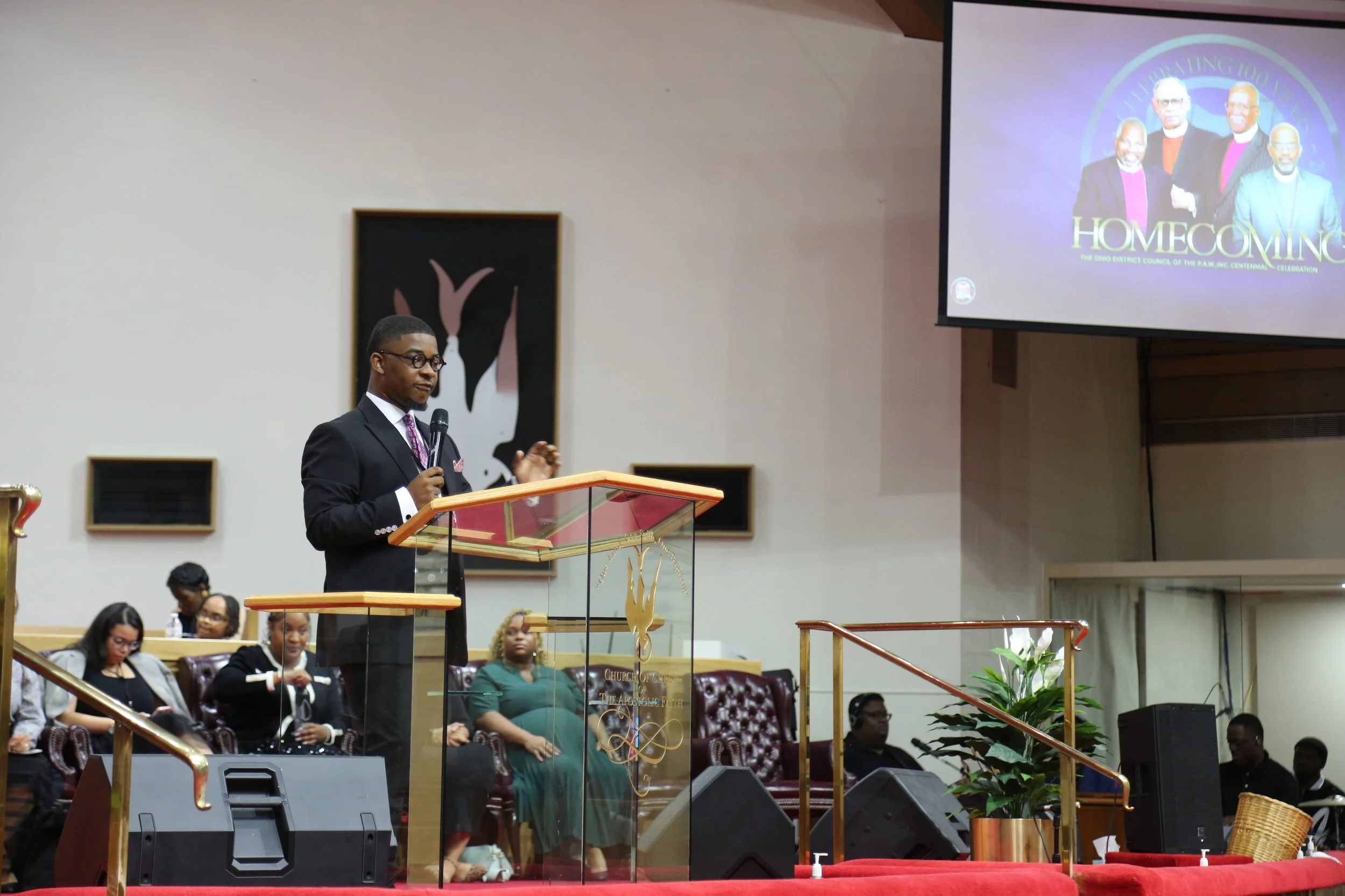 A young man in a dark suit and glasses speaking at a church podium with a microphone. Behind him, seated people including women, some with head coverings, and a large screen displaying a group of men in suits with the word 'HOMECOMING'.