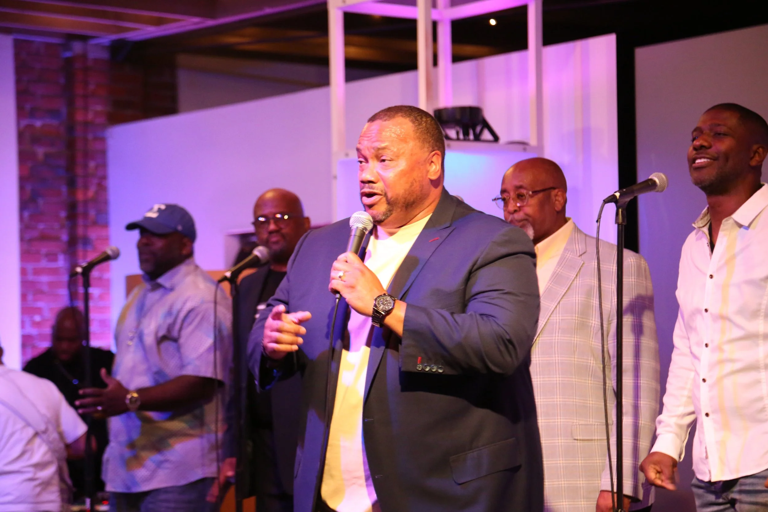 A group of five men, four standing and one sitting, on a stage with microphones, at an indoor event with purple lighting and brick wall background. The man in front is speaking into a microphone, wearing a dark blazer and a watch.