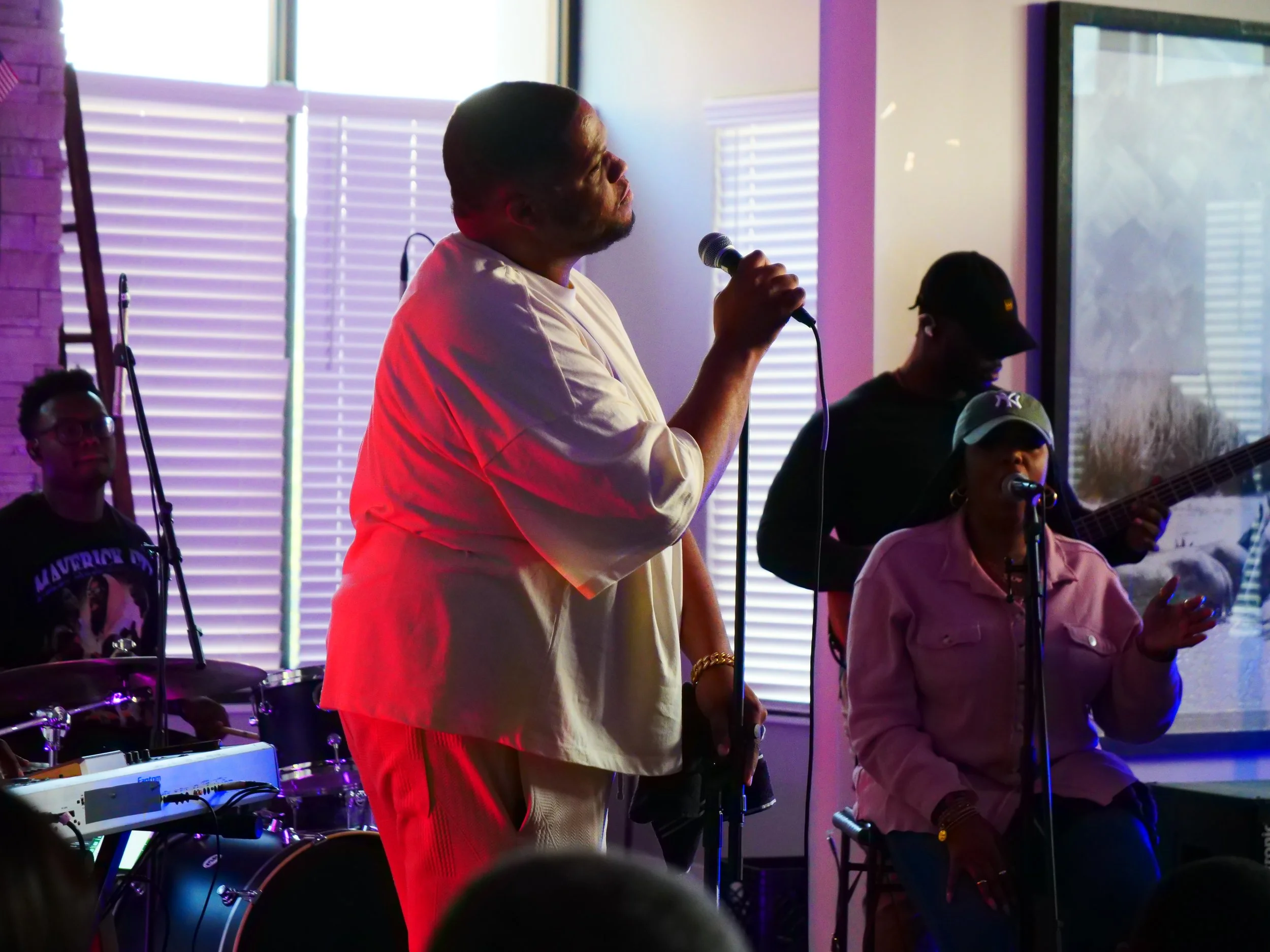 A band performing indoors with a male singer holding a microphone, a female singer seated with a microphone, a guitarist, and a drummer in the background, illuminated by colorful lighting near window blinds.