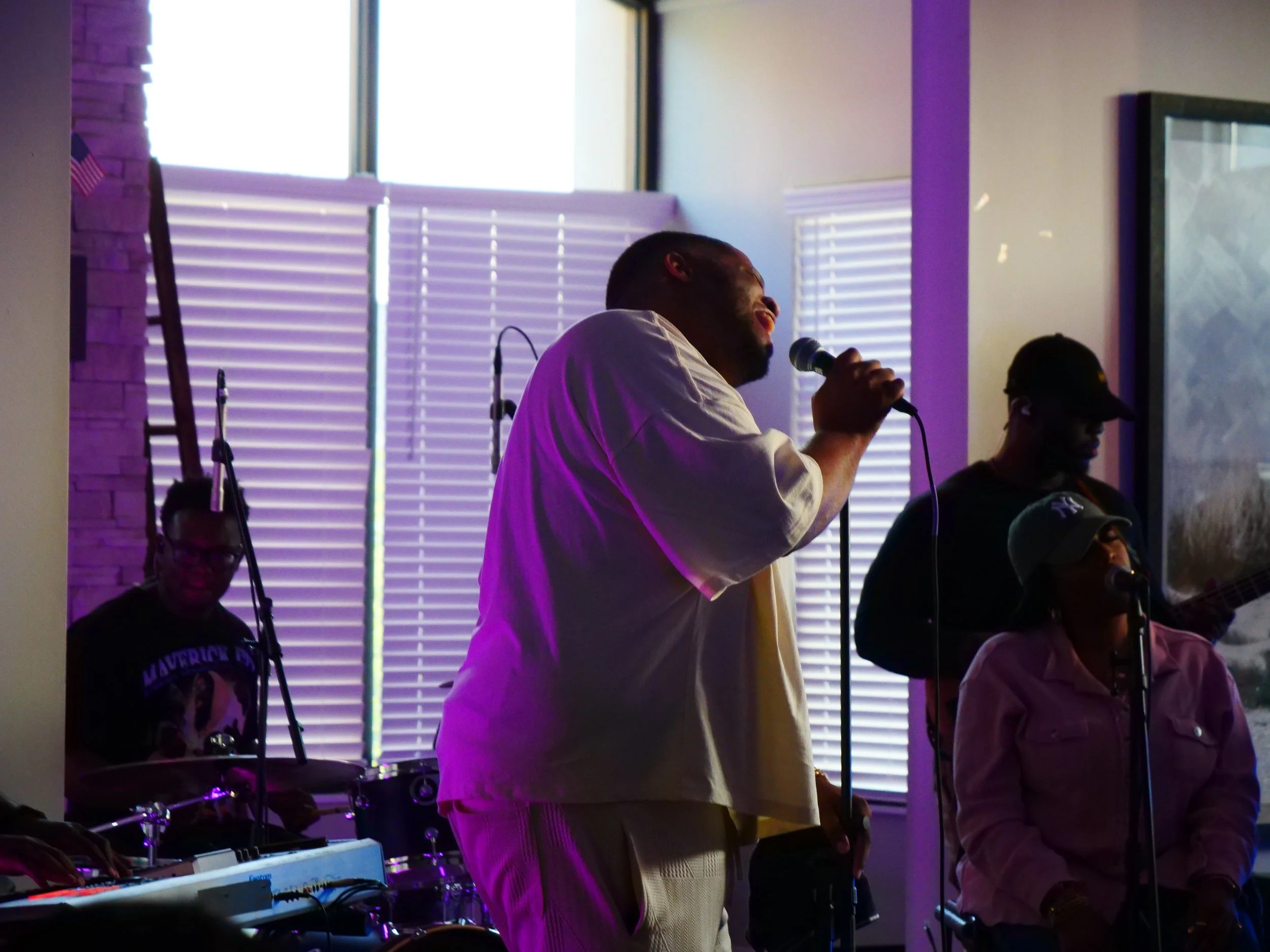A musician singing into a microphone with a band playing behind him, indoors with window blinds in the background.