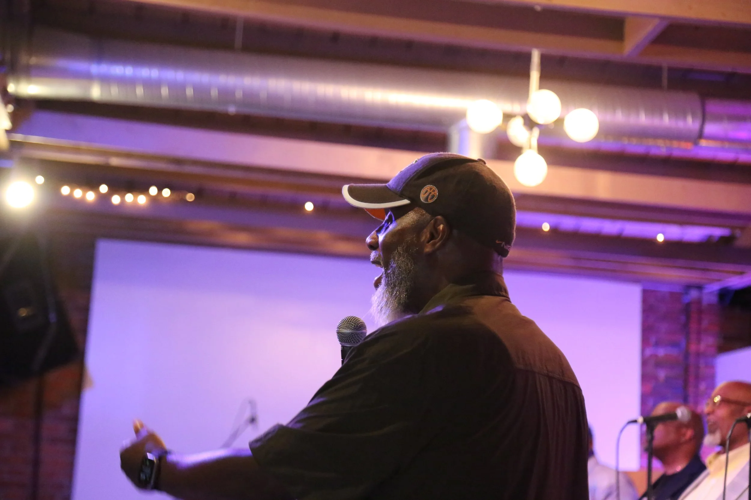 A man with a beard wearing a baseball cap and sunglasses, holding a microphone, standing in a dimly lit venue with purple lighting and hanging globe lights.