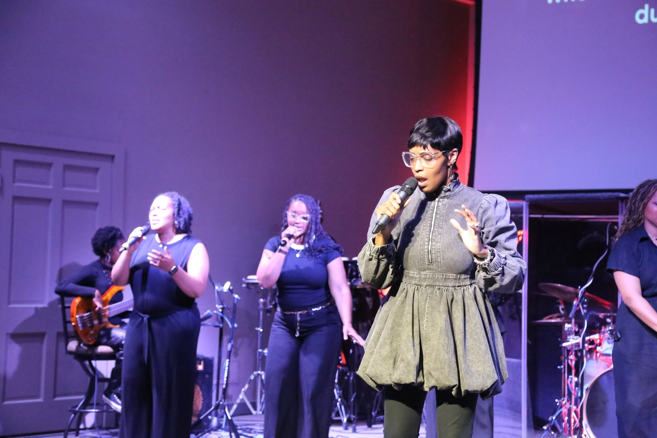 A group of women singing and playing musical instruments on stage, with a backdrop and large screen in the background.