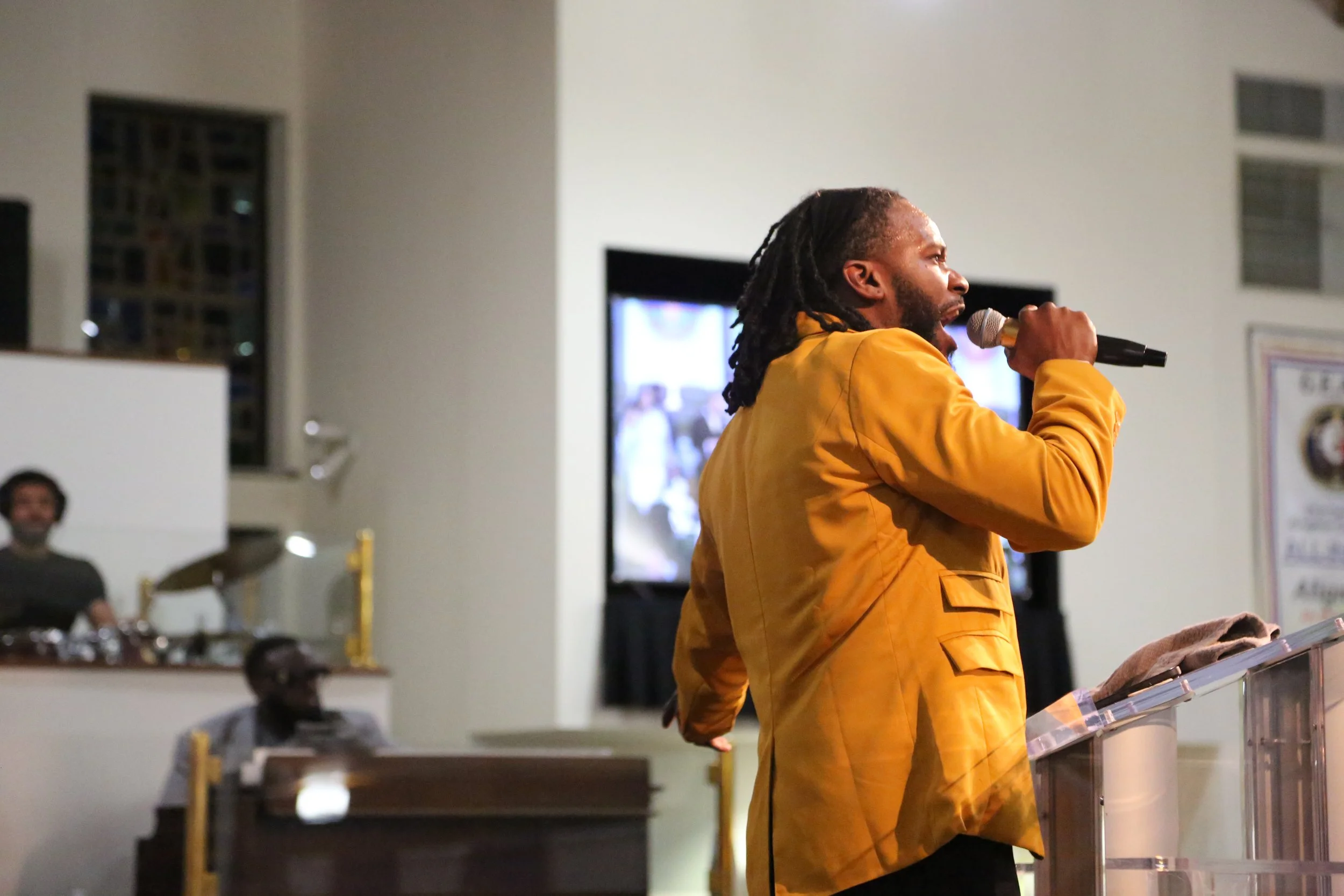 A man with dreadlocks and a beard wearing a yellow jacket speaking or singing into a microphone at a podium in a room with multiple people present.