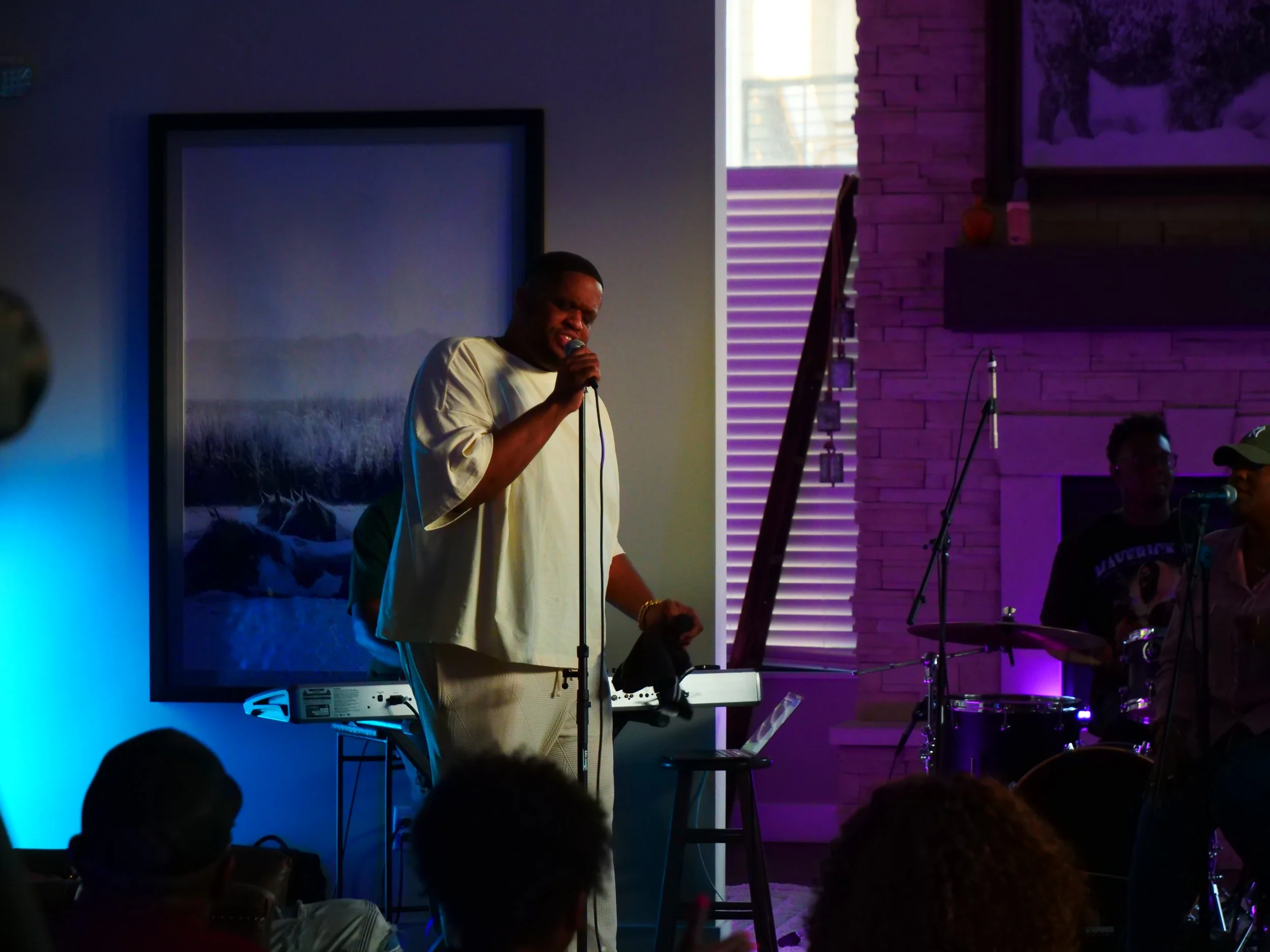 A man singing into a microphone during a live performance with musical instruments and audience members in a dimly lit venue.