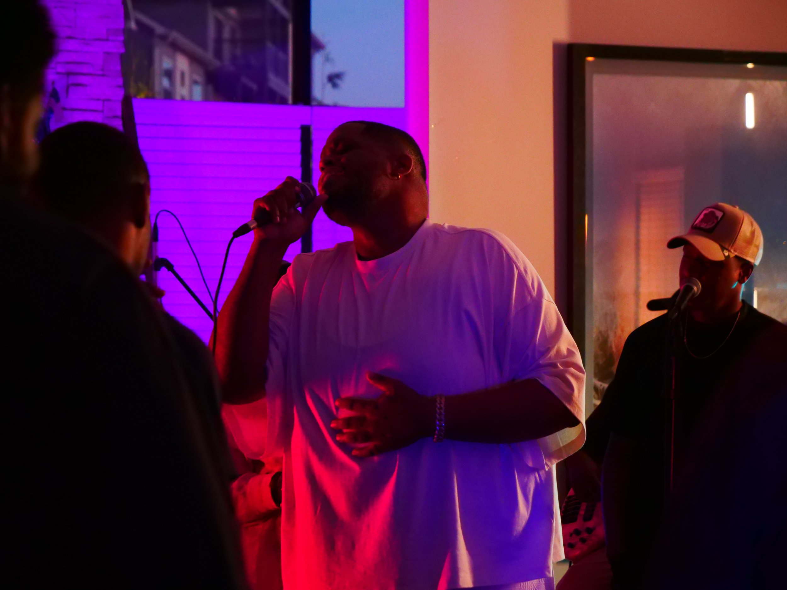 A man in a white shirt singing into a microphone in a dimly lit room with purple and orange lighting, with two other men nearby, one wearing a cap and what appears to be a black shirt.