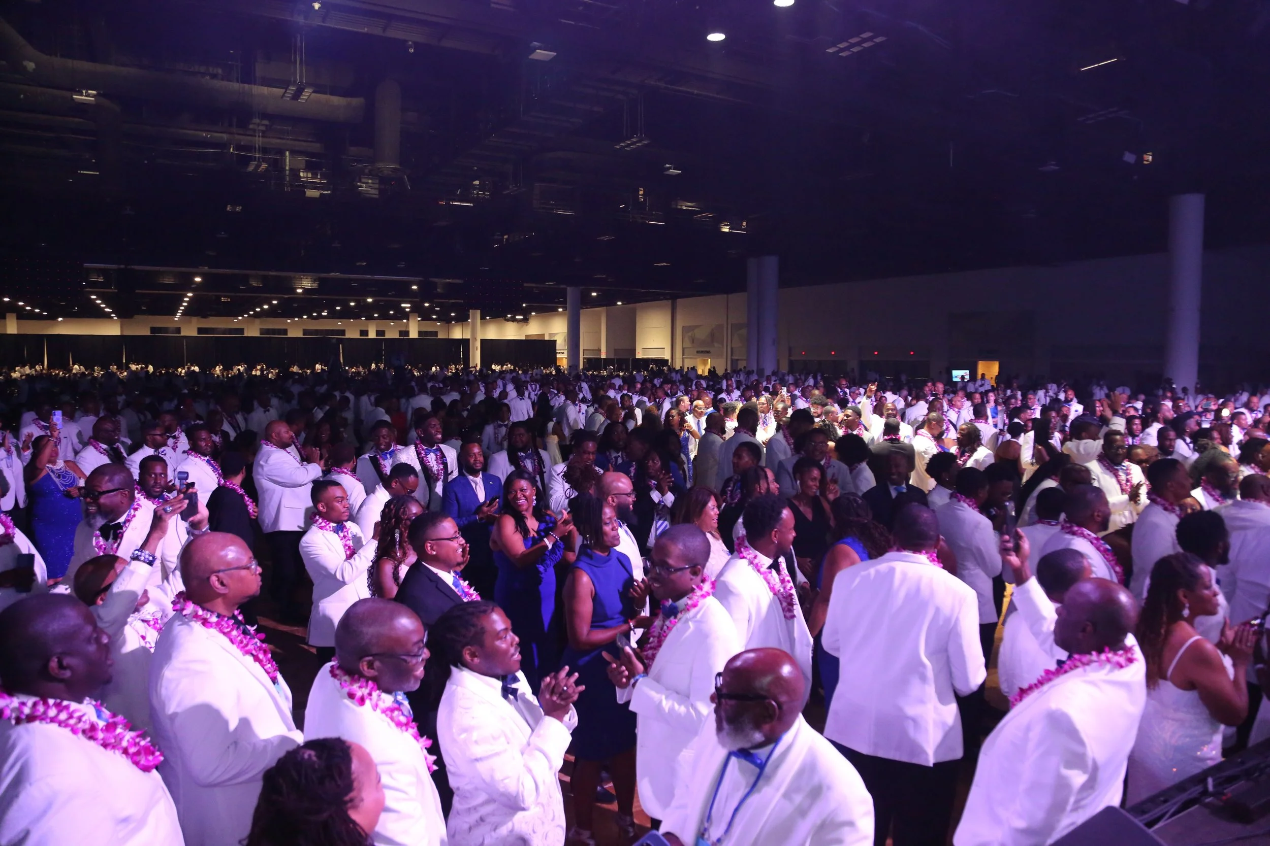 Large indoor gathering of people dressed in formal attire, many wearing leis, in a spacious, dimly lit venue with purple lighting.