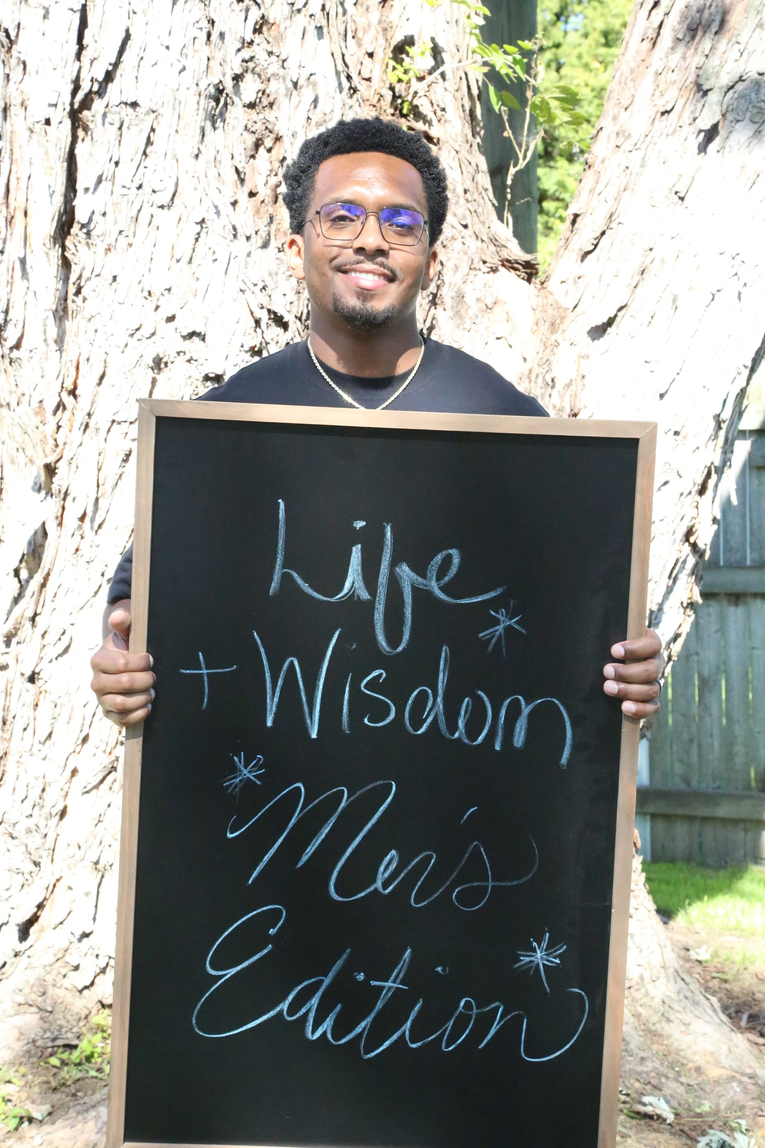 A smiling man with glasses and a necklace holding a blackboard with the words 'Hope + Wisdom = Men's Edits' written on it, standing outdoors in front of a large tree.
