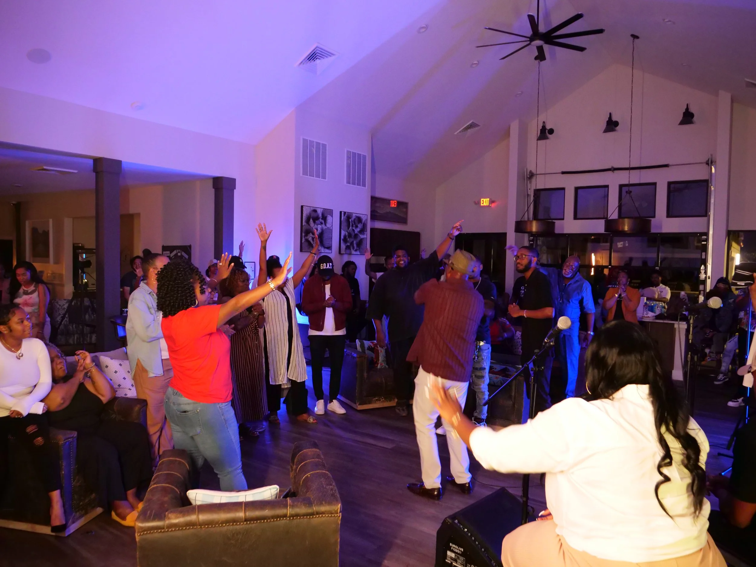 People attending a lively social gathering or party in a spacious, well-lit indoor venue with artwork on the walls, some with hands raised, engaging in music and dancing.