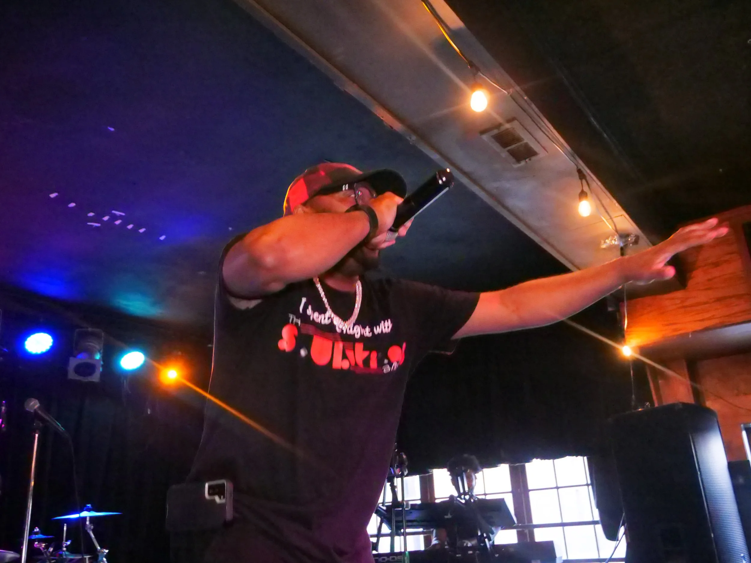 A man wearing a black t-shirt, glasses, and a red cap is performing on stage, holding a microphone in one hand and extending his other arm outward, in a dimly lit venue with colored stage lights.