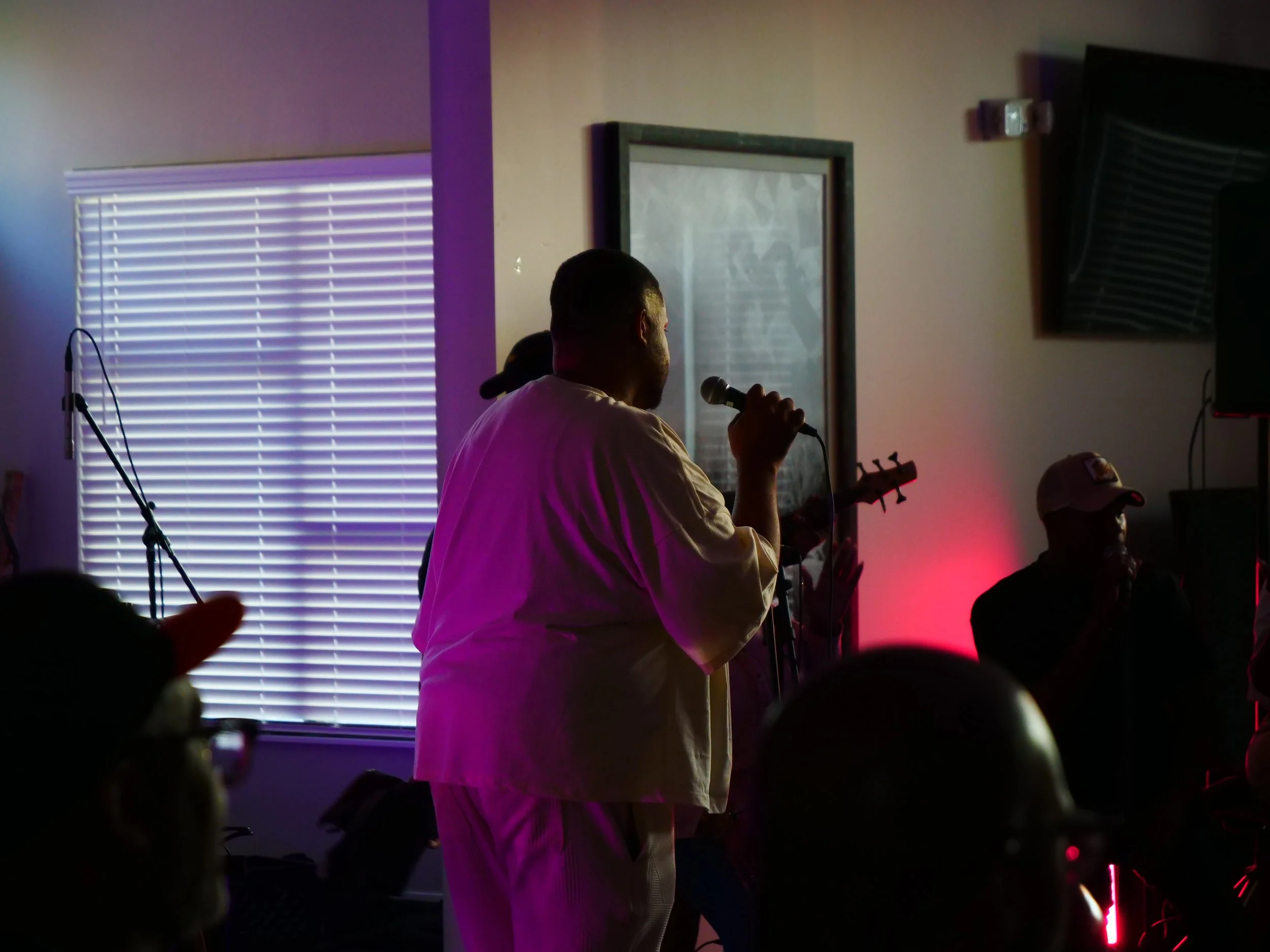 A person holding a microphone and playing an electric guitar during a performance, with a band behind him, in a dimly lit room with colored lighting.