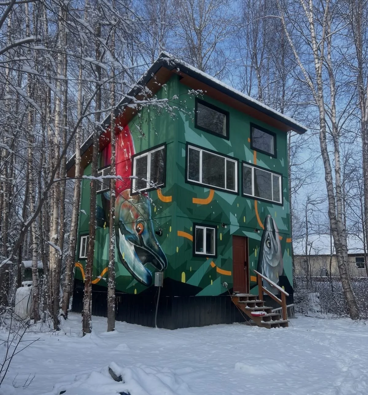 A colorful house with a mural of a fish and a bird in a snowy forest. The house has multiple windows, a wooden staircase, and a green background with abstract shapes and lines.