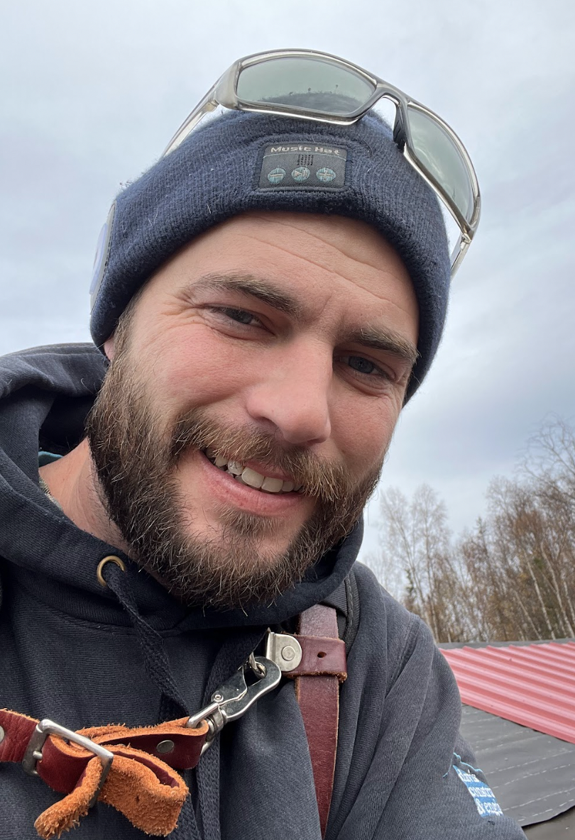 A man outdoors smiling, wearing a gray beanie with sunglasses on top, and a dark hoodie. The background shows leafless trees and a red and black metal roof.