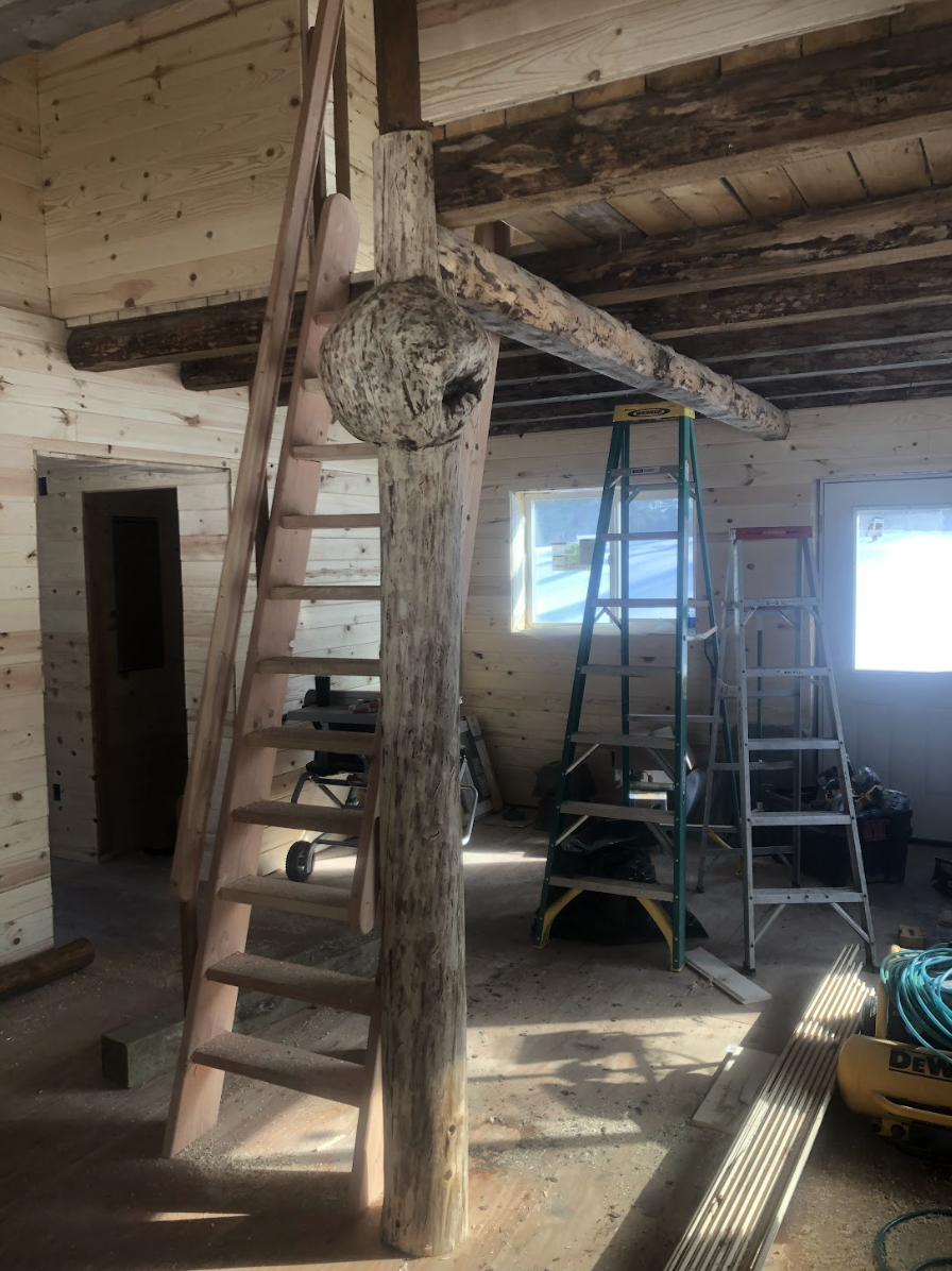 Interior of a building under construction with wooden walls and ceiling, featuring a rustic wood support beam, a wooden ladder leaning against the beam, and several stepladders, along with construction tools and materials scattered on the floor.